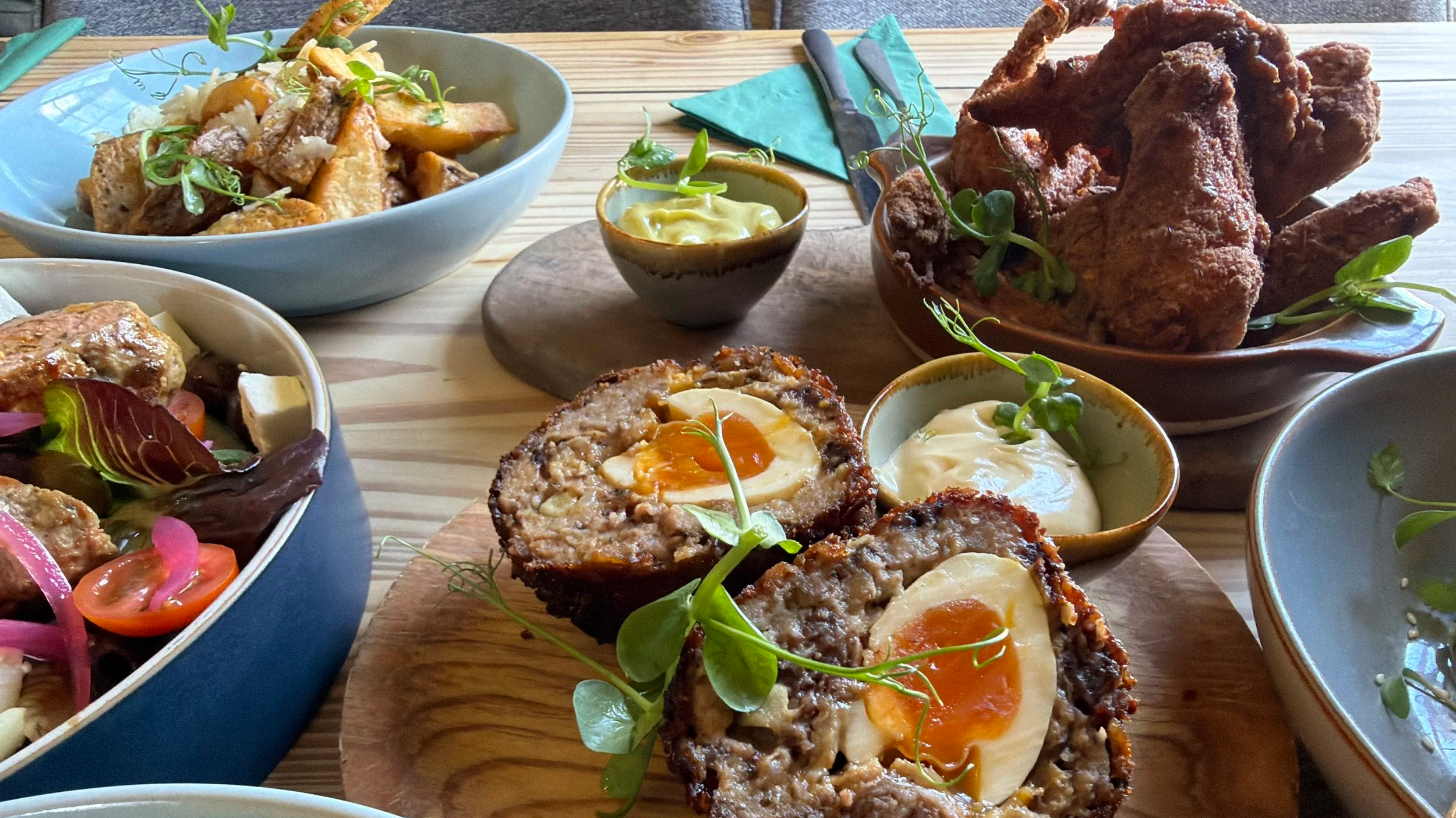 Various plates of food on a wooden table, including baked eggs in a meatload, a salad, roasted chicken, and bowls of sauces, garnished with microgreens.