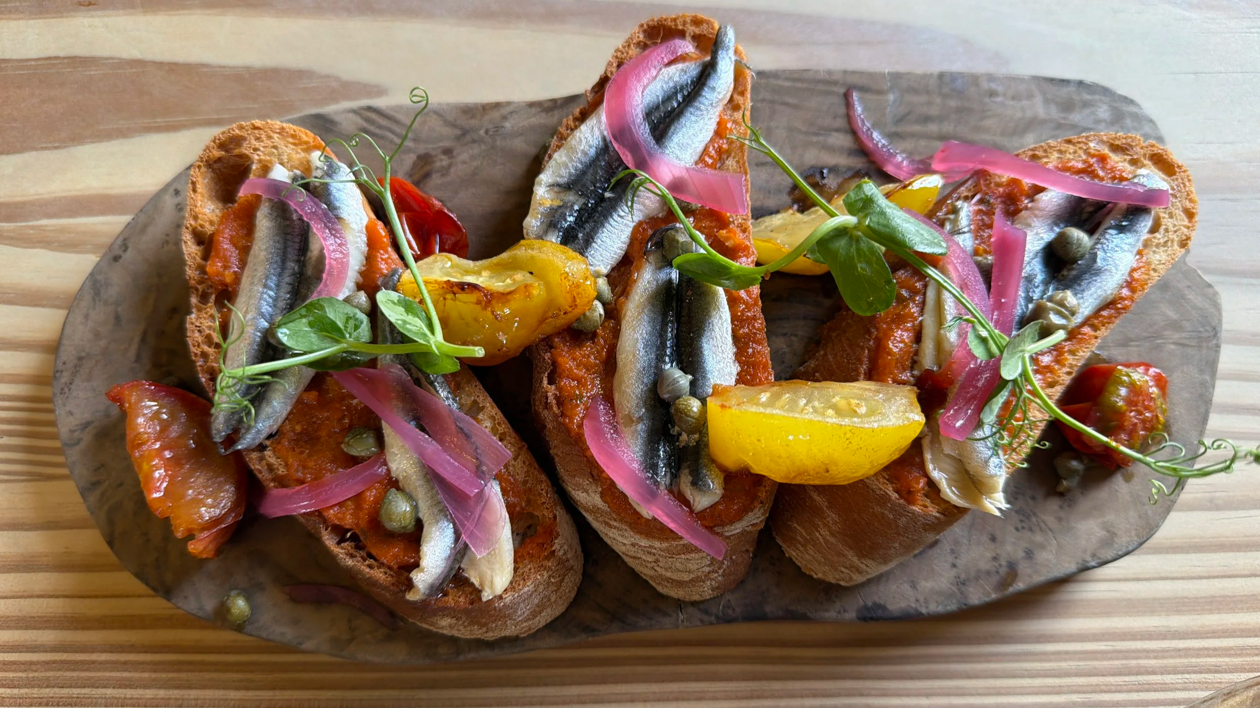 Open-faced sandwiches with sardines, cherry tomatoes, pickled onions, roasted yellow peppers, capers, and fresh greens served on a wooden platter.