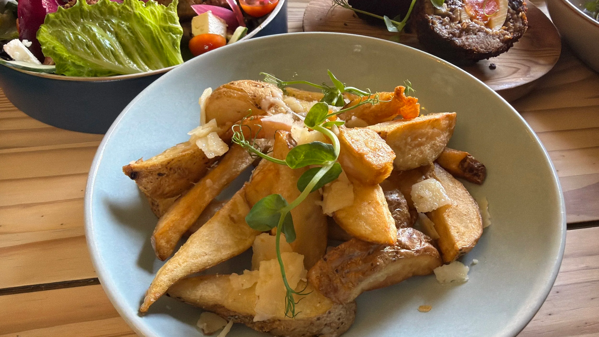 Bowl of roasted potato wedges with garnishing on top, with side salads and stuffed mushroom in the background on wooden table.