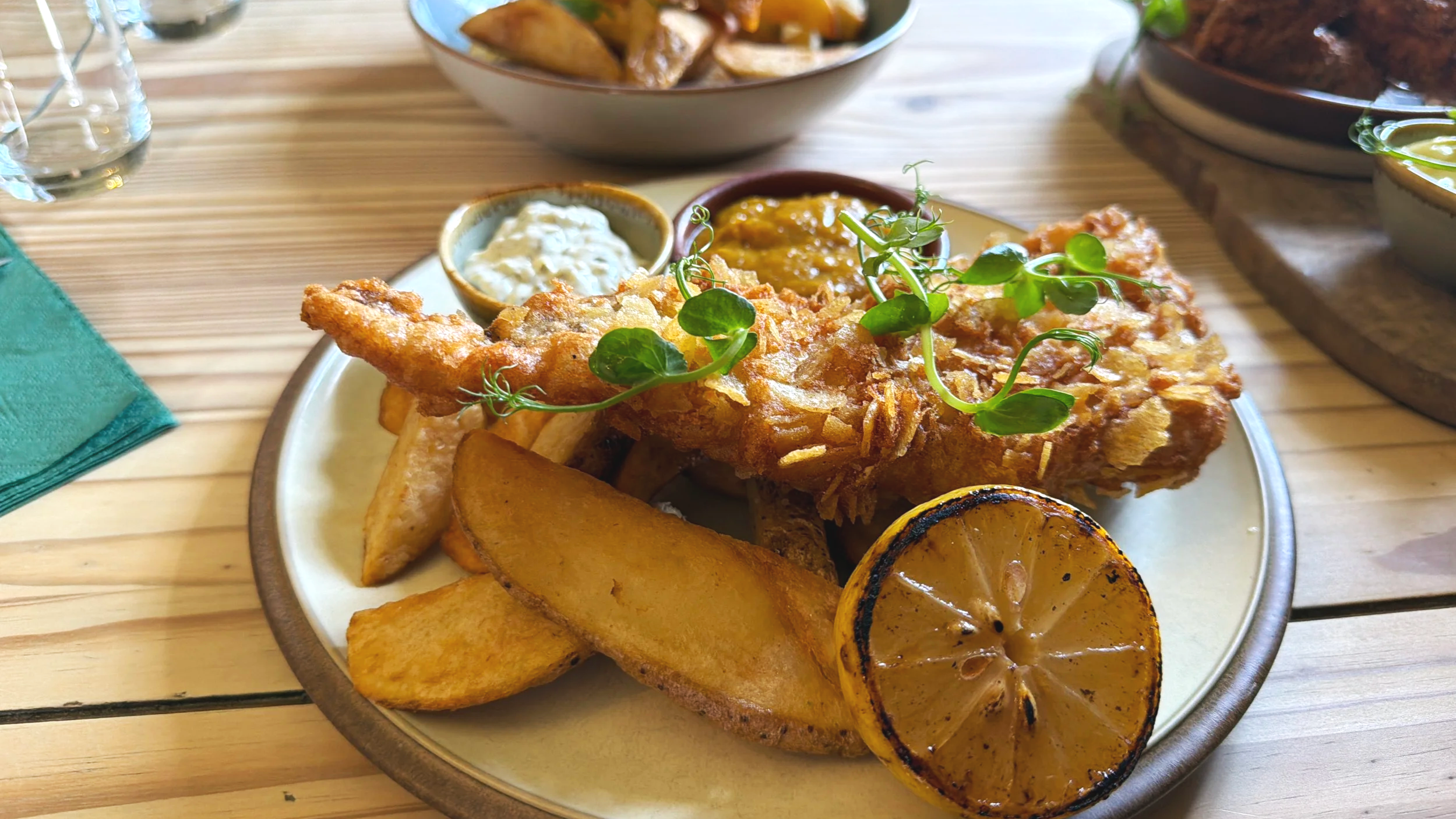 Fried fish with lemon wedge, grilled potatoes, potato wedges, tartar sauce, and baked beans on a plate, garnished with greens.