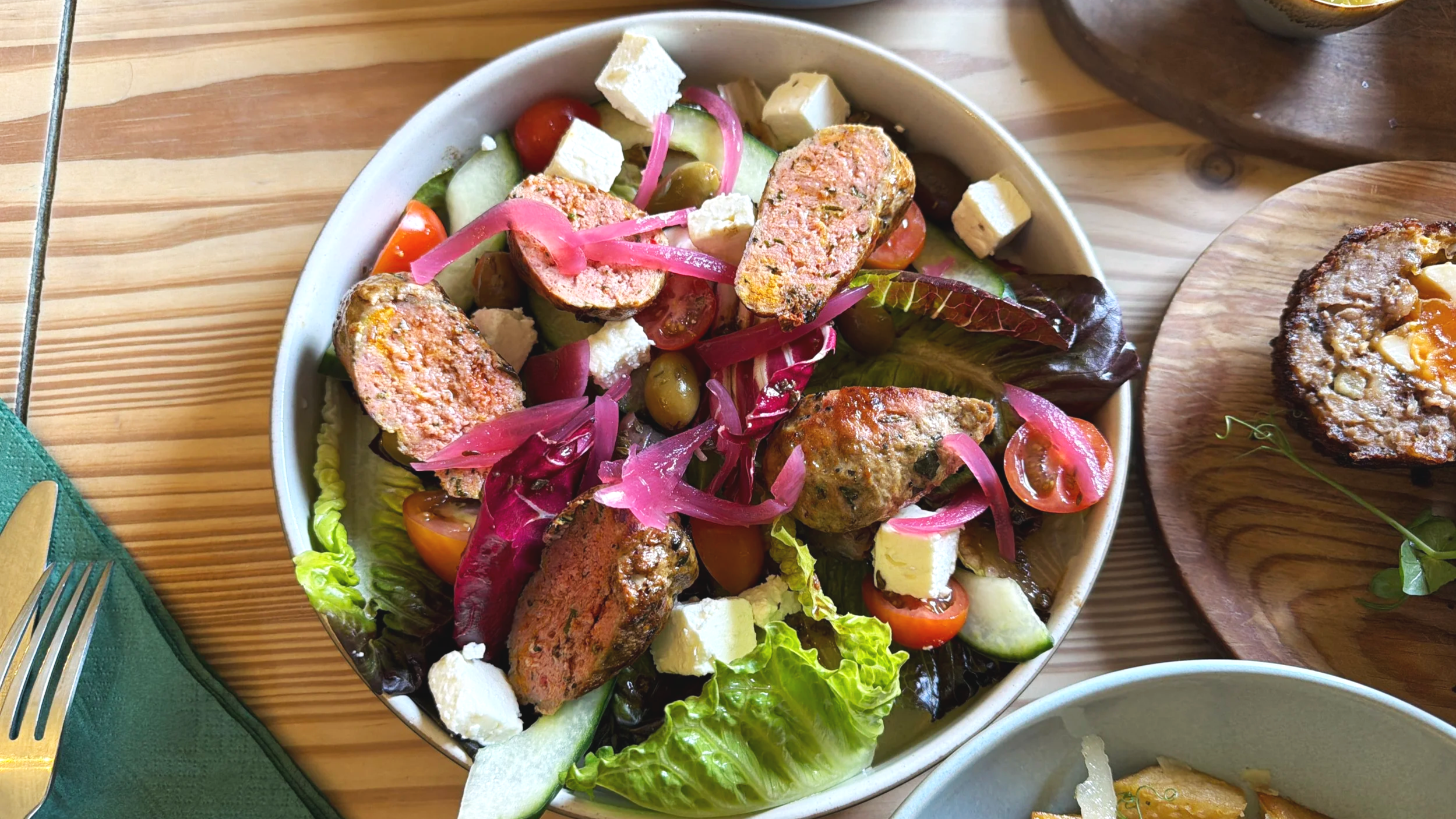 A bowl of mixed salad containing cherry tomatoes, cucumbers, olives, feta cheese, pickled onions, and sliced pieces of seasoned grilled chicken, on a wooden table with a green napkin, fork, and other dishes nearby.