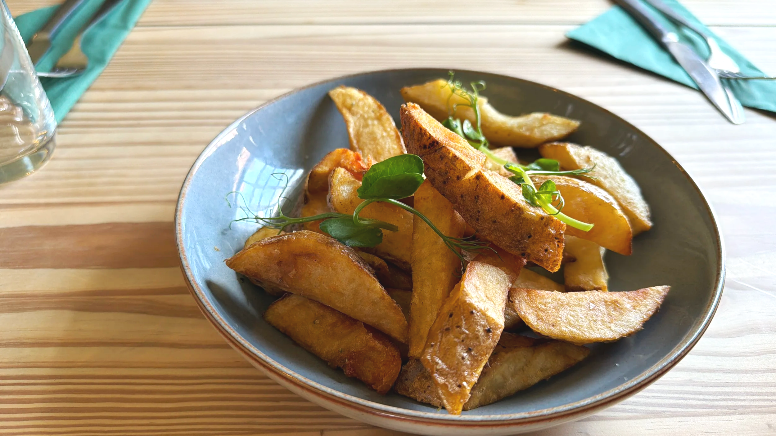 A bowl of roasted potato wedges garnished with fresh green herbs on a wooden table