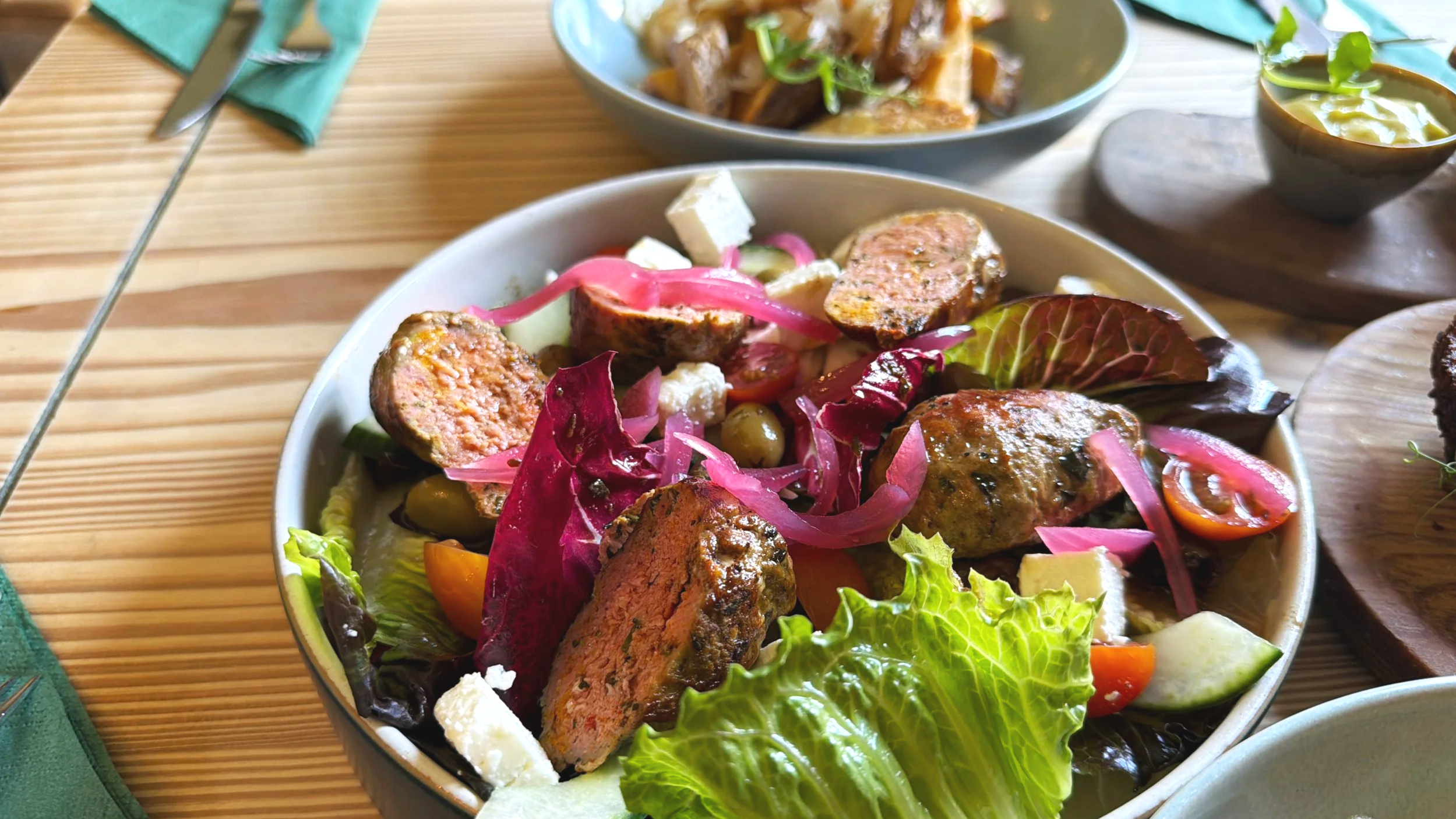 Salad with sliced sausage, lettuce, tomato, pickles, feta cheese, and pickled red onions in a bowl on a wooden table.