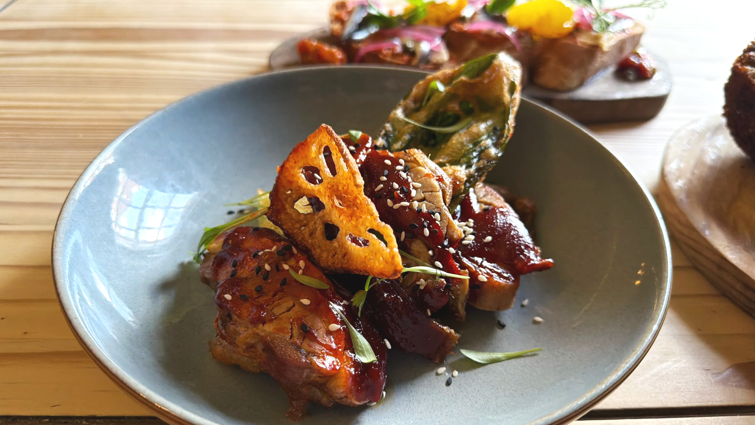 Close-up of a gourmet appetizer with seared meat, garnished with sesame seeds, microgreens, and crispy lotus root chip, served on a light blue plate.