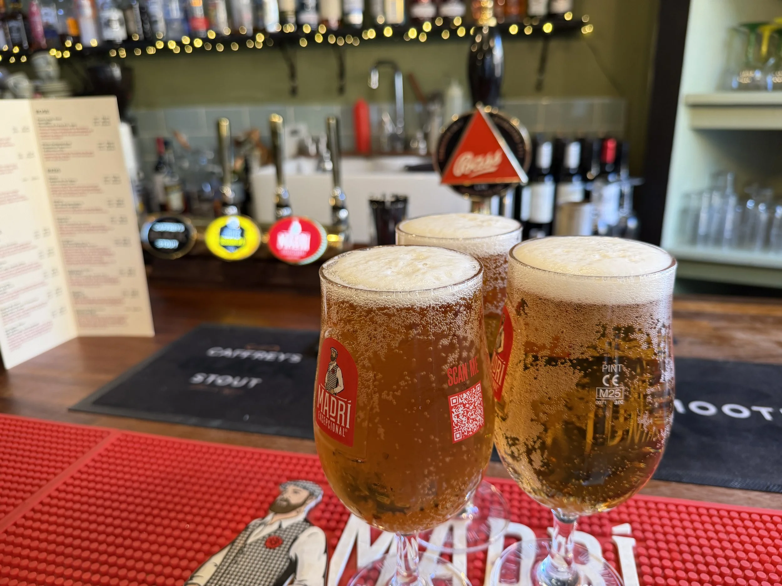 Three glasses of beer on a bar counter with a menu and bar equipment in the background.