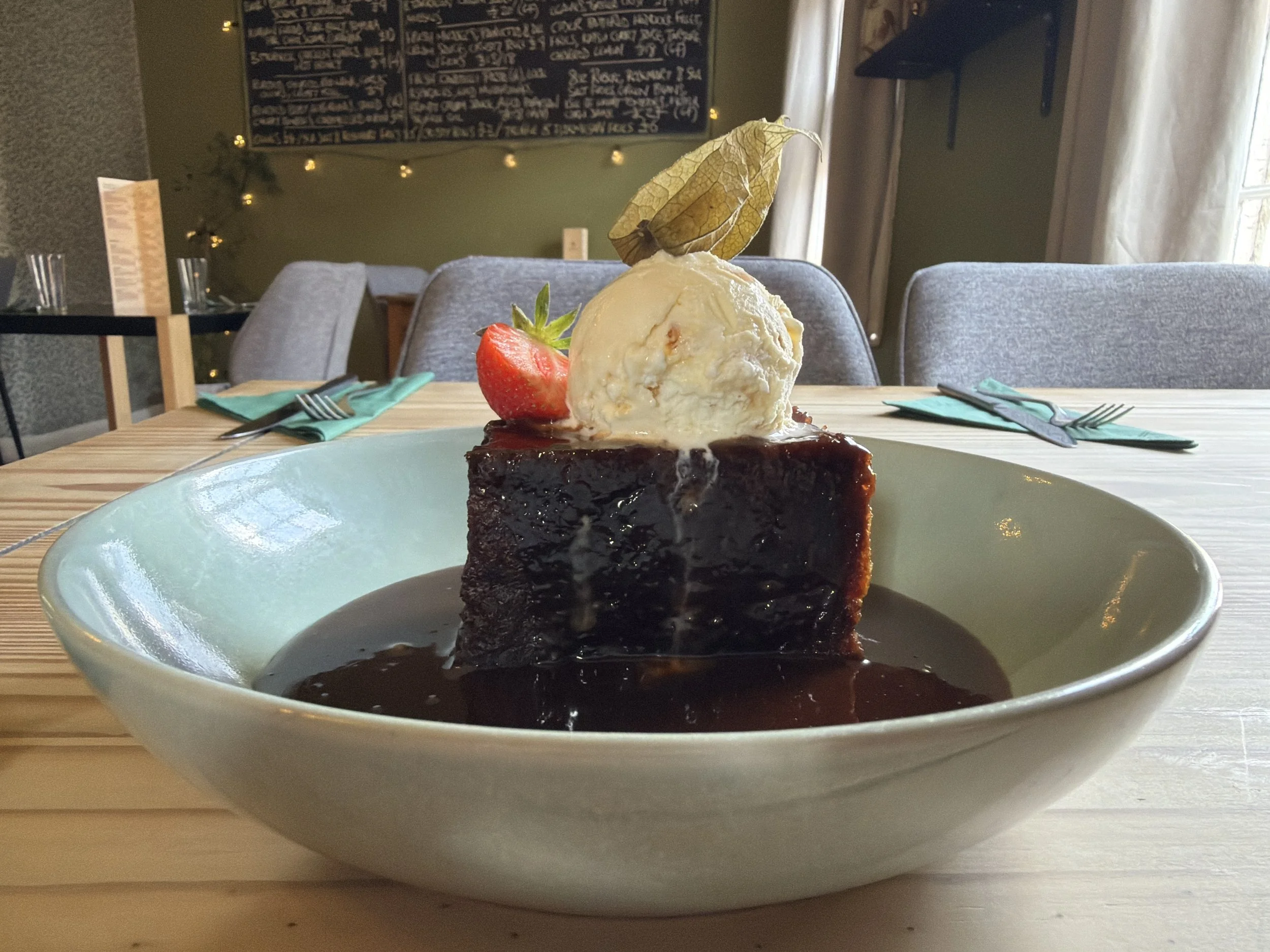 A dessert featuring a brownie topped with a scoop of vanilla ice cream, a strawberry, and a dried physalis on a light blue plate, set on a wooden table in a restaurant.