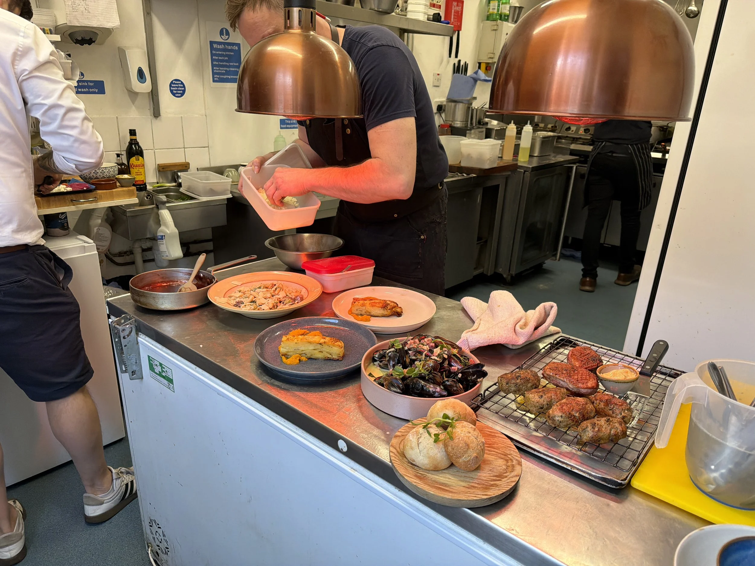 A busy kitchen with chefs preparing and serving various dishes, including bread and baked items, under copper heat lamps.