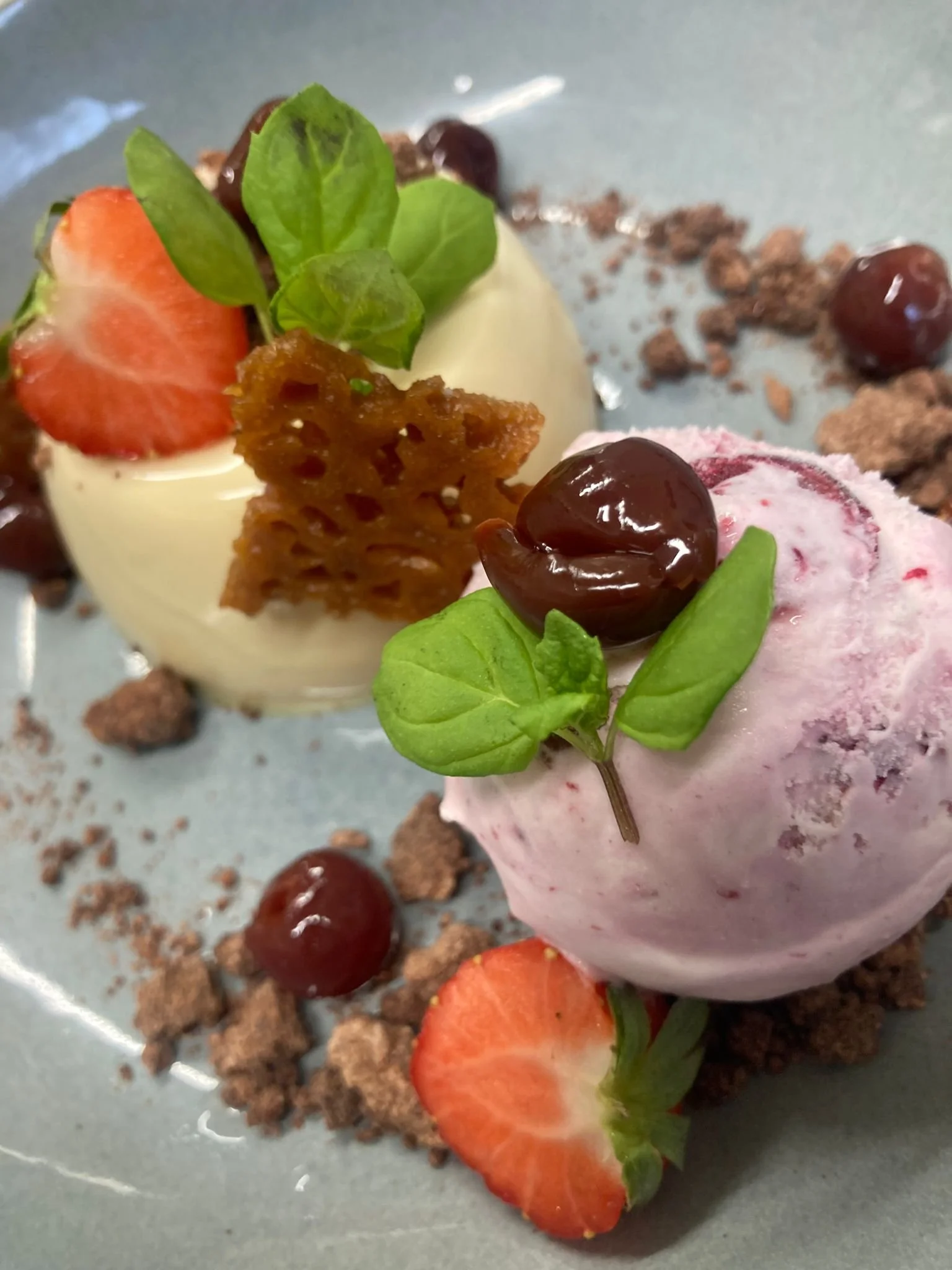Close-up of a dessert with sliced strawberries, blueberries, fresh mint leaves, a scoop of pink ice cream, a white chocolate piece, and a crispy caramel lattice, garnished with chocolate crumbles and scattered berries on a gray surface.