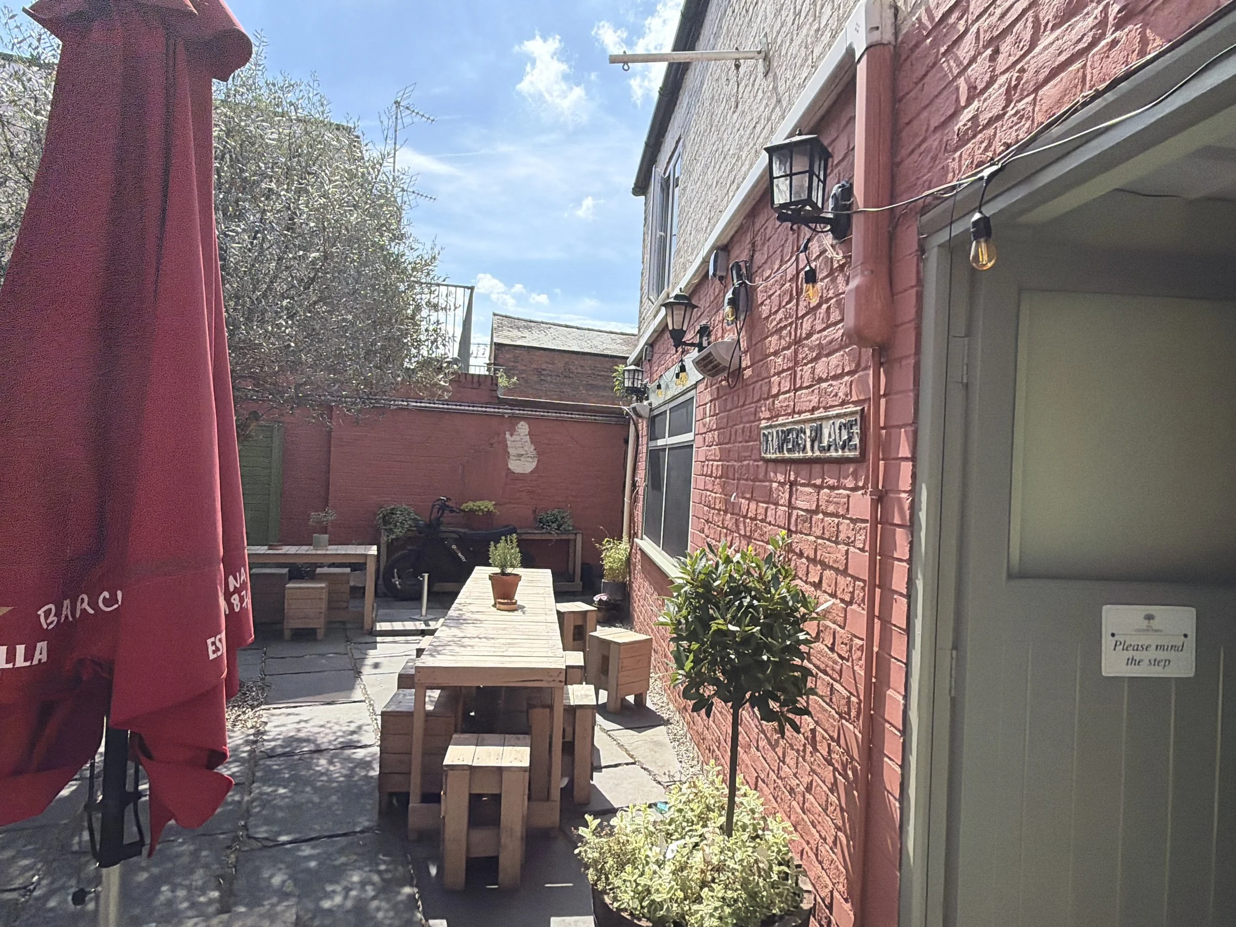 Outdoor patio area with red brick wall, wooden tables and chairs, potted plants, and a red patio umbrella. The sky is clear and sunny.