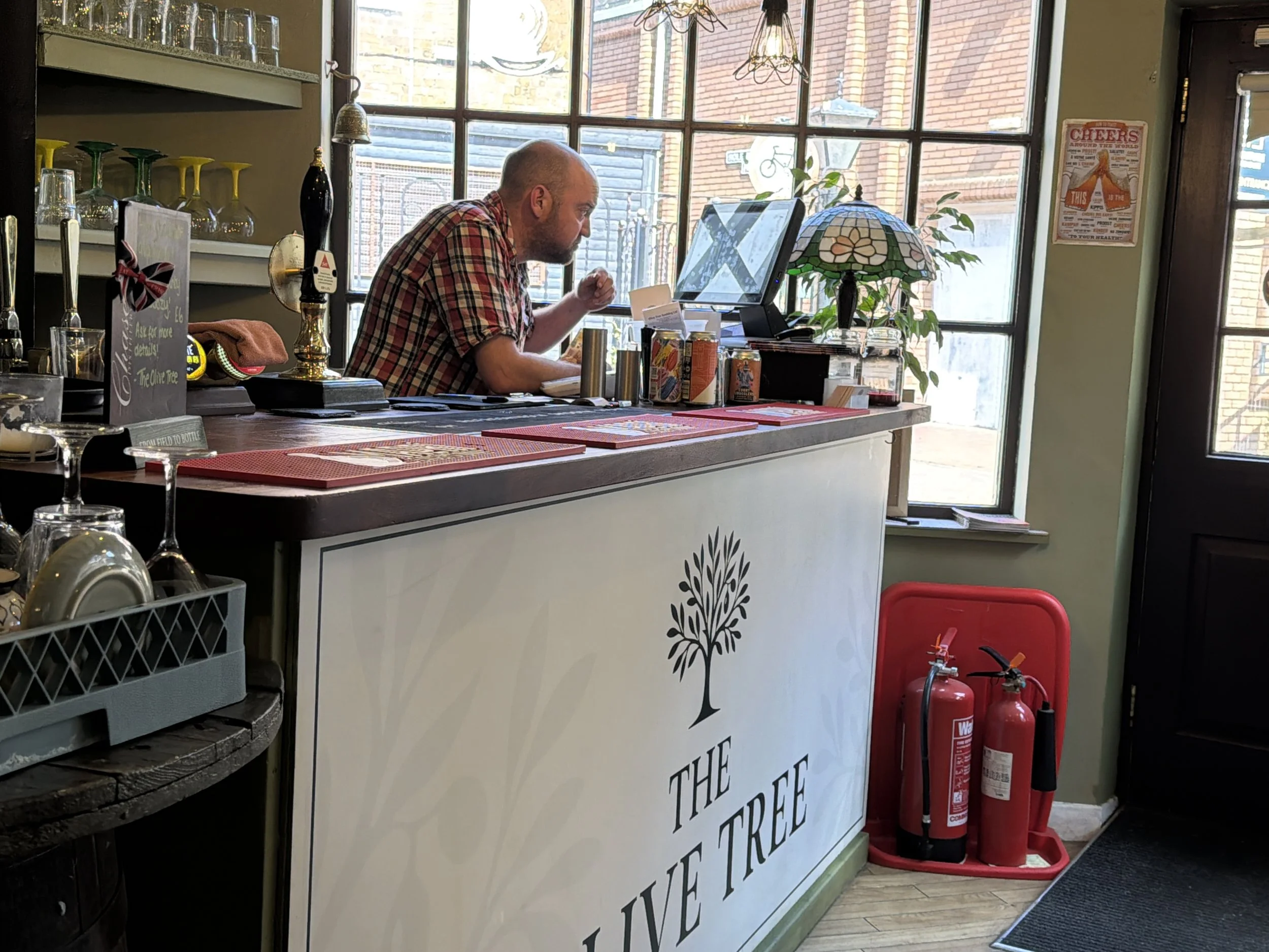 A man in a plaid shirt working at a counter in a café or restaurant with a large window behind him. There are drinks, a lamp, and a computer on the counter. Fire extinguishers are visible near the door on the right.