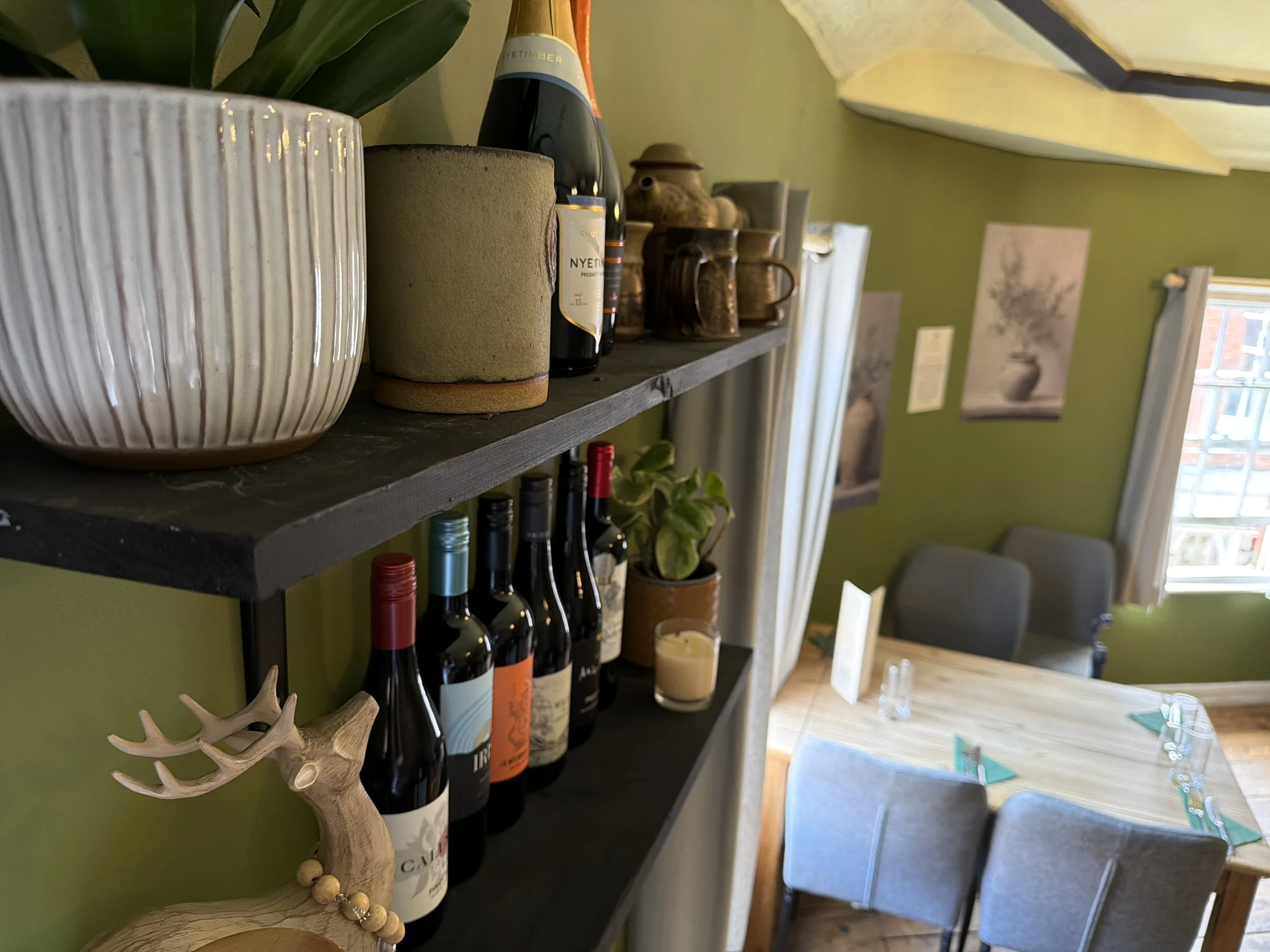 A close-up of a black shelf holding wine bottles, a potted plant, empty ceramic cups, and decorative items in a cozy dining room with green walls and artwork.
