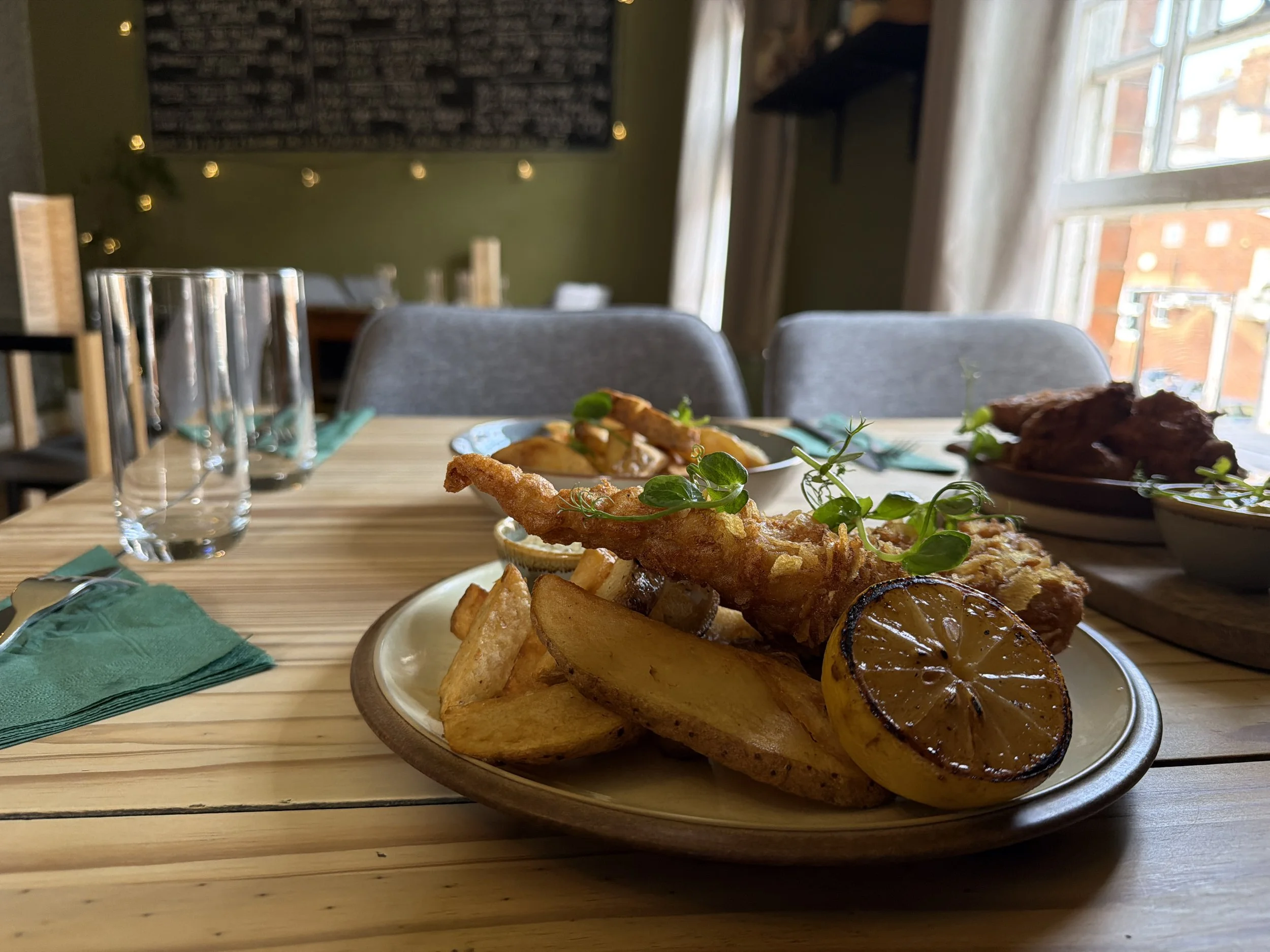 A wooden table with a plate of roasted vegetables including grilled lemon slices, and fried fish fillets garnished with herbs. In the background, there are empty glasses, green napkins, and a dining room with green walls, curtains, and large windows 