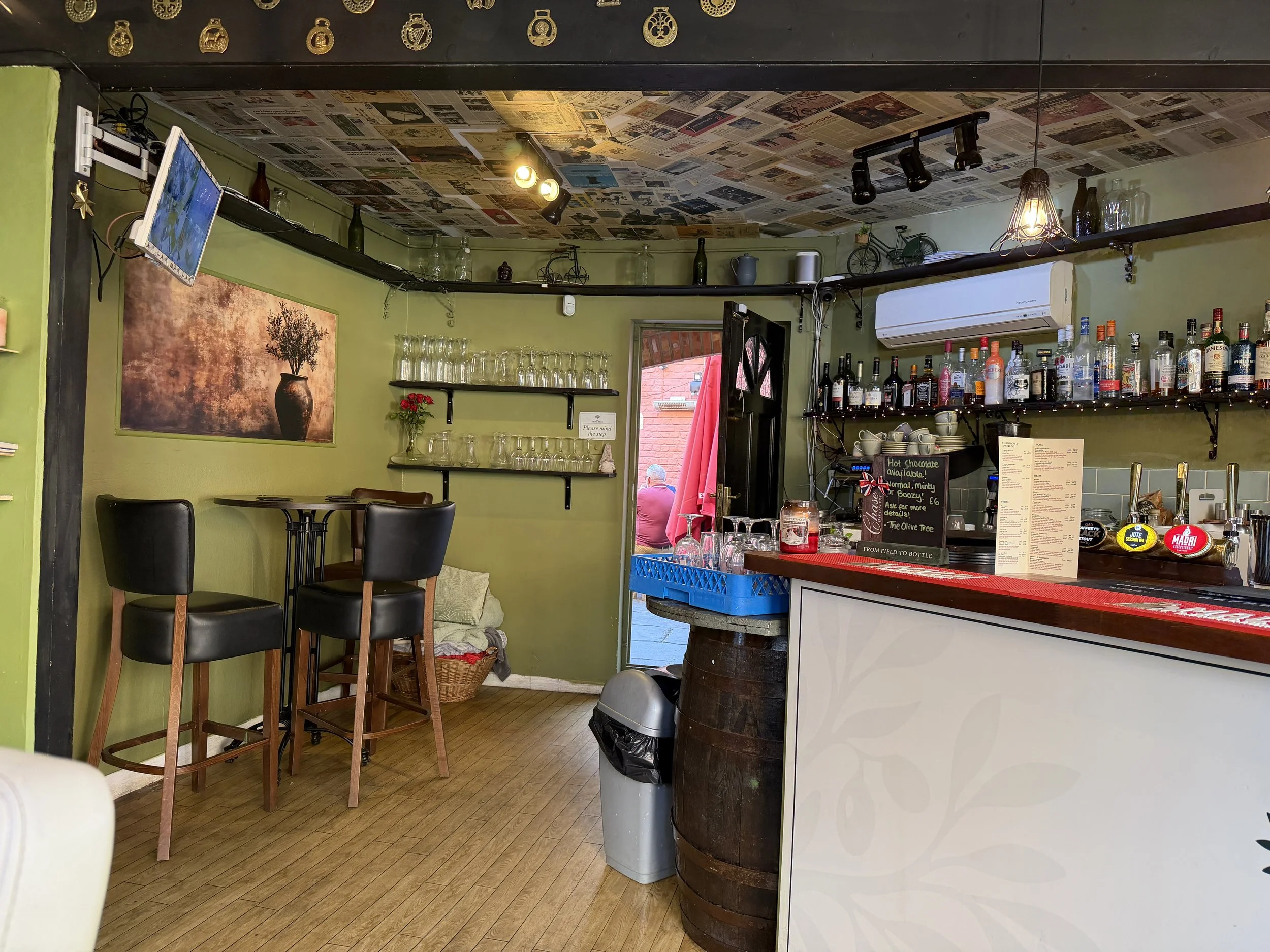 The interior of a cozy cafe with green walls, decorated with newspaper pages on the ceiling, shelves with glassware, bottles, and decorative items, a chalkboard menu on a barrel, and a small dining area with chairs and a painting of a plant.