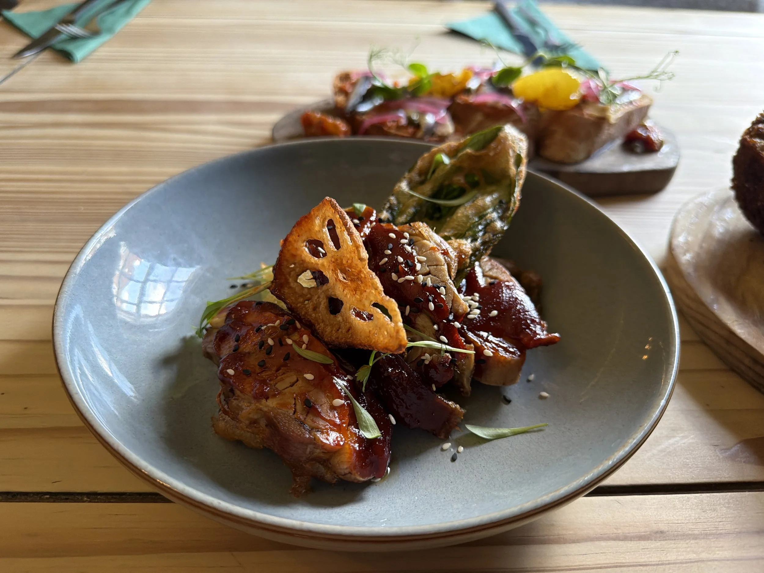 Close-up of a bowl of sushi with salmon, topped with sesame seeds, green herbs, and a sweet sauce. In the background, there are various sushi pieces garnished with herbs and toppings on a wooden table.