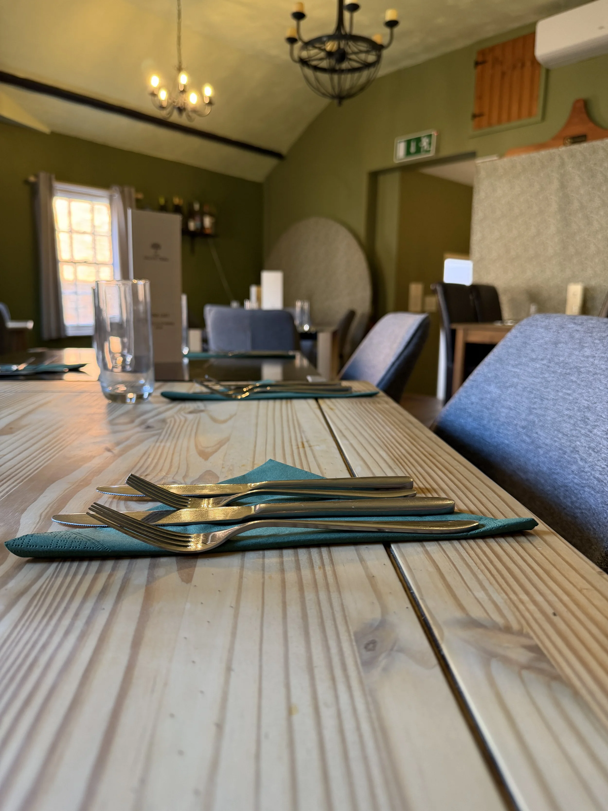 Table setting in a restaurant with forks, knives, and teal napkins placed on a light wooden table.