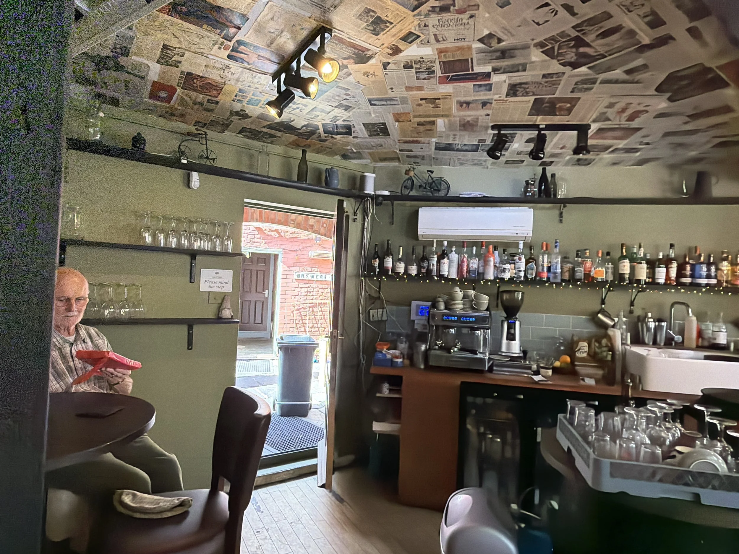 Interior of a cozy cafe or restaurant with a newspaper-covered ceiling, a bar area with liquor bottles, glasses, and kitchen equipment, a man sitting at a table, and an open door revealing an outdoor area.