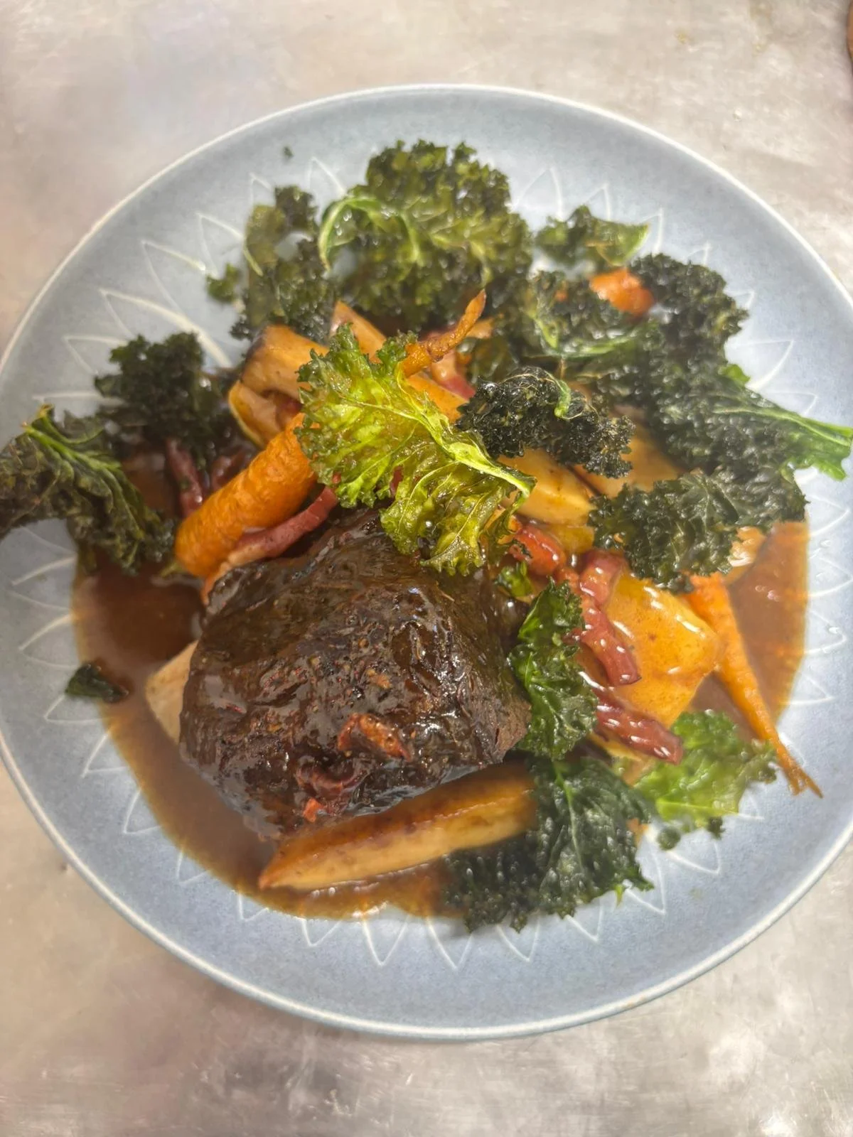 A plated dish featuring a cooked whole fish with dark sauce, surrounded by kale, carrots, potatoes, and shredded meat in a brown sauce.