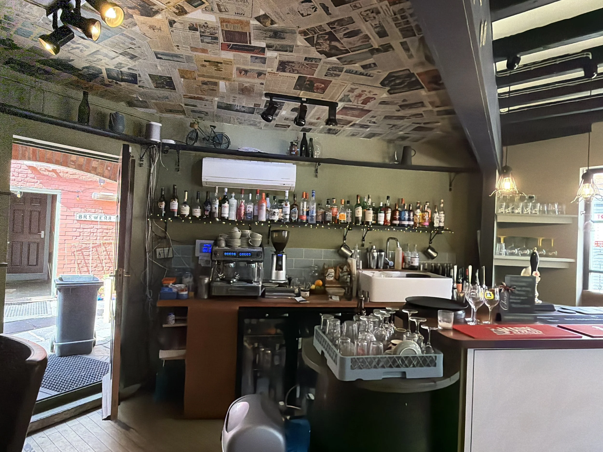 Interior of a cozy bar or cafe with a newspaper-covered ceiling, shelves with bottles and cups, a window, and dim lighting.