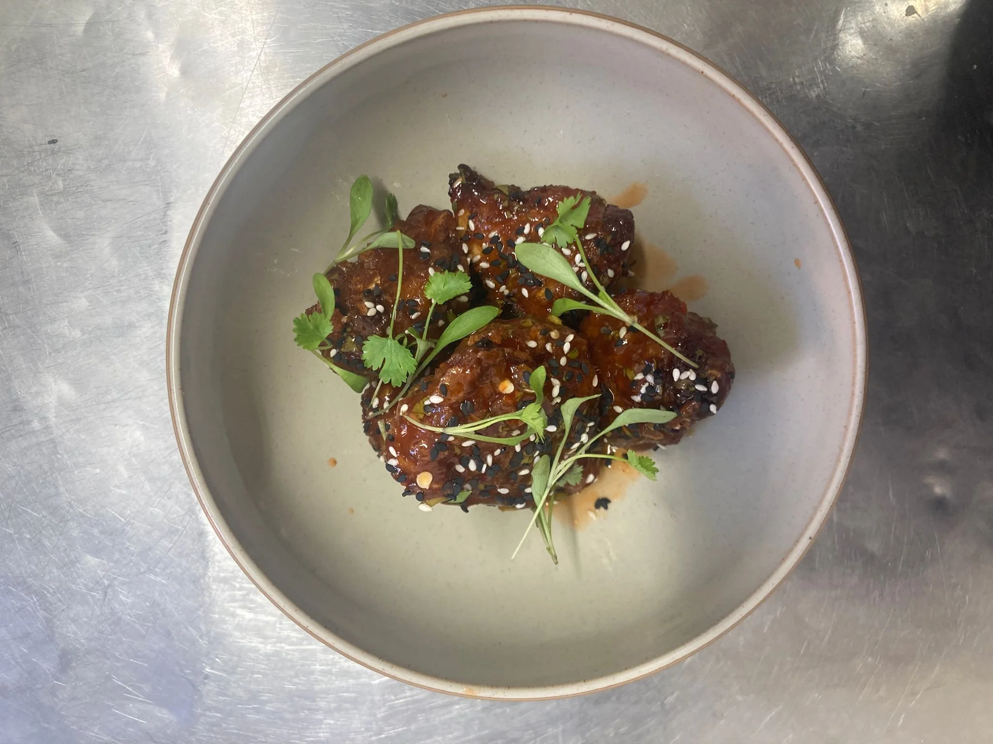 A white bowl with a dish of cooked meat, garnished with fresh green herbs and sprinkled with black and white sesame seeds.