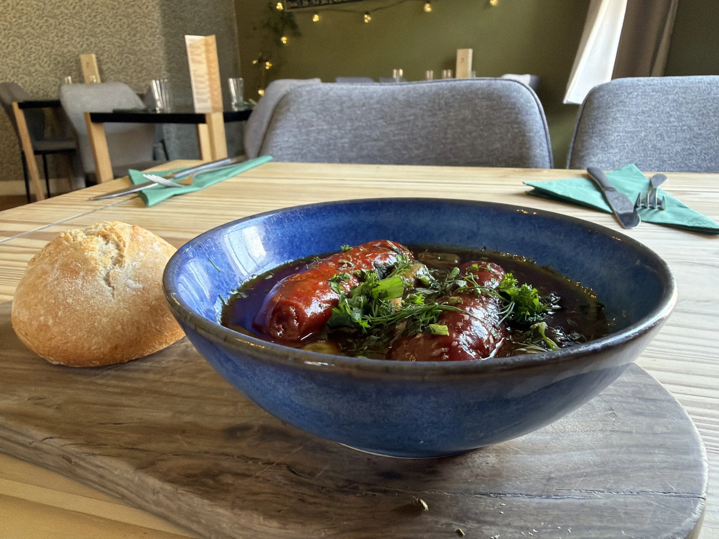 A blue bowl of stuffed tomatoes with herbs, a piece of bread on a wooden table, set in a restaurant with gray chairs and fairy lights in the background.
