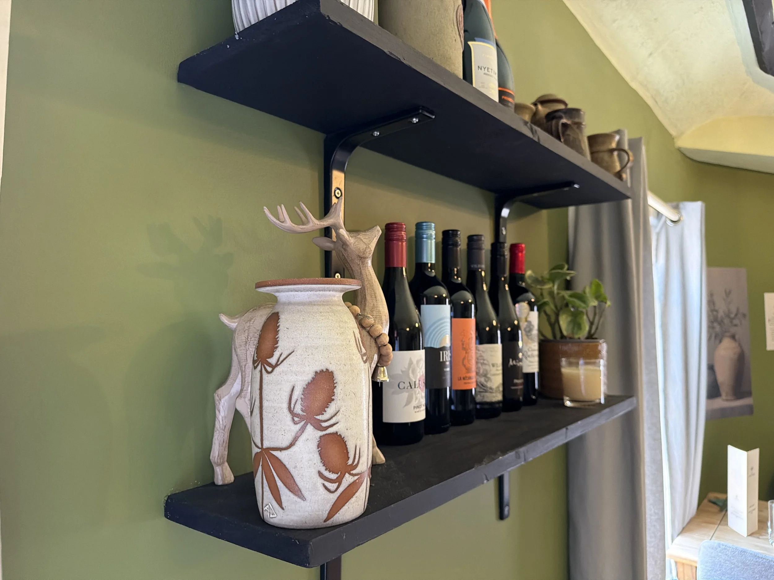 Decorative ceramic vase with antler-inspired design on a wall-mounted black shelf, surrounded by wine bottles and other decorative items in a home interior with a green wall.
