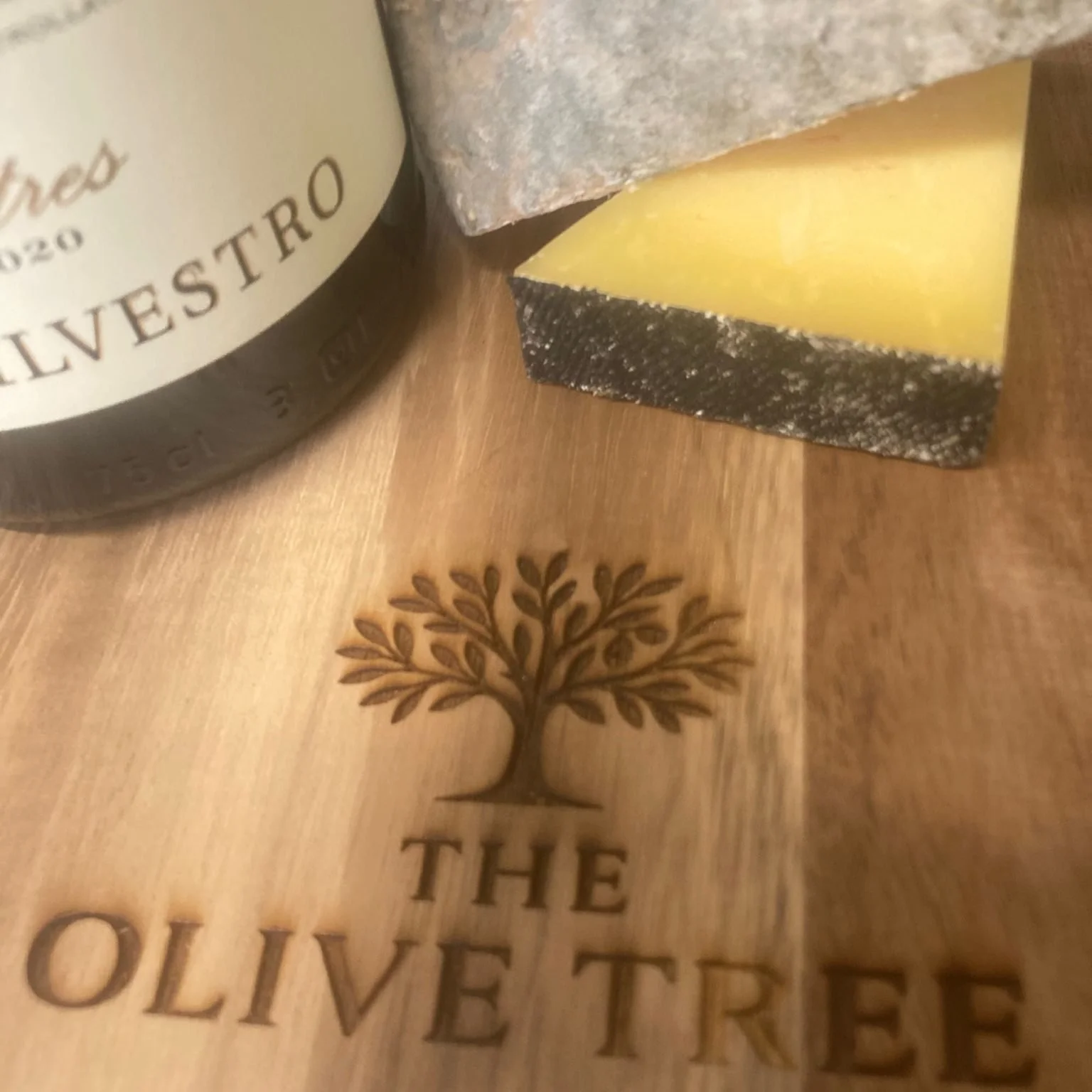 A wooden surface with a decorative tree engraving and carved text, partially showing the words 'THE OLIVE TREE'. A wedge of cheese and a grayish rectangular stone or bread are also on the surface.