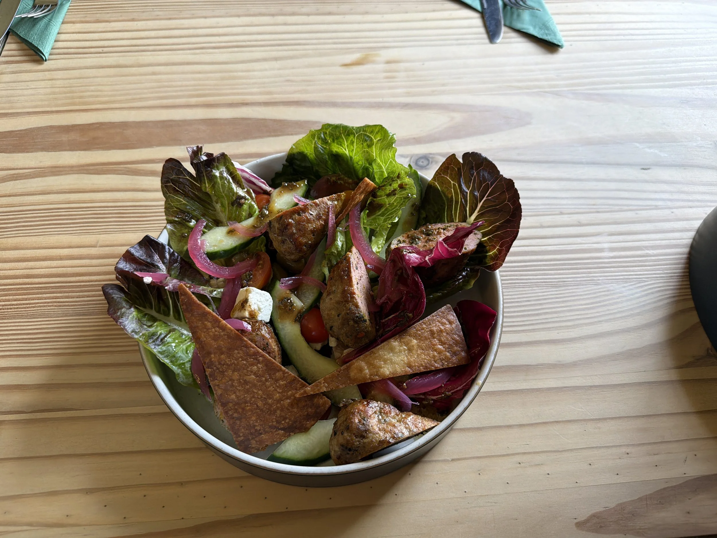 A bowl of grilled chicken, pita bread, salad with lettuce, onion, cucumber, and cherry tomatoes, on a wooden table.