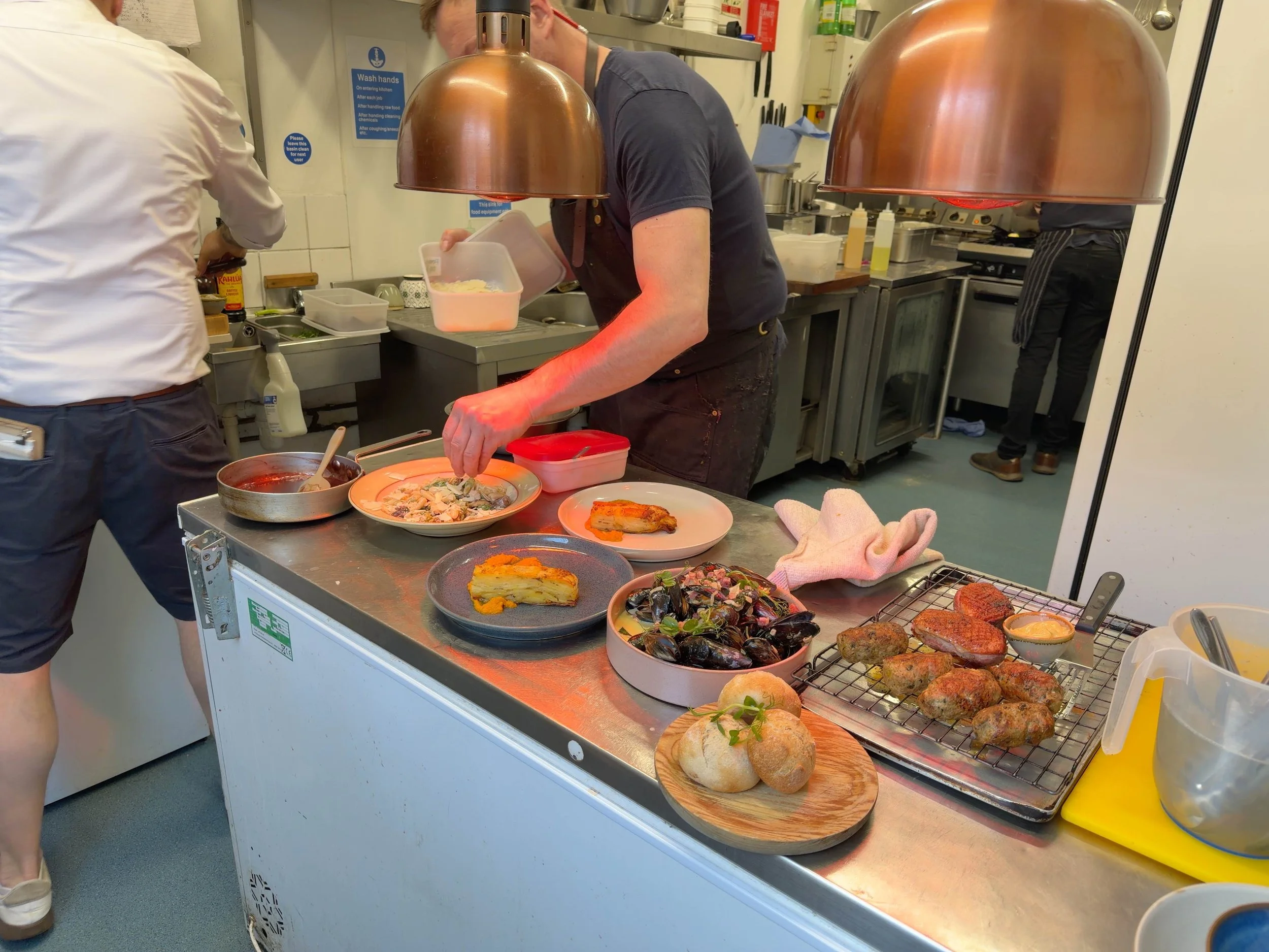 A busy kitchen with chefs preparing and plating dishes, featuring a countertop with various food items including bread, salads, and fried food, under copper pendant lights.