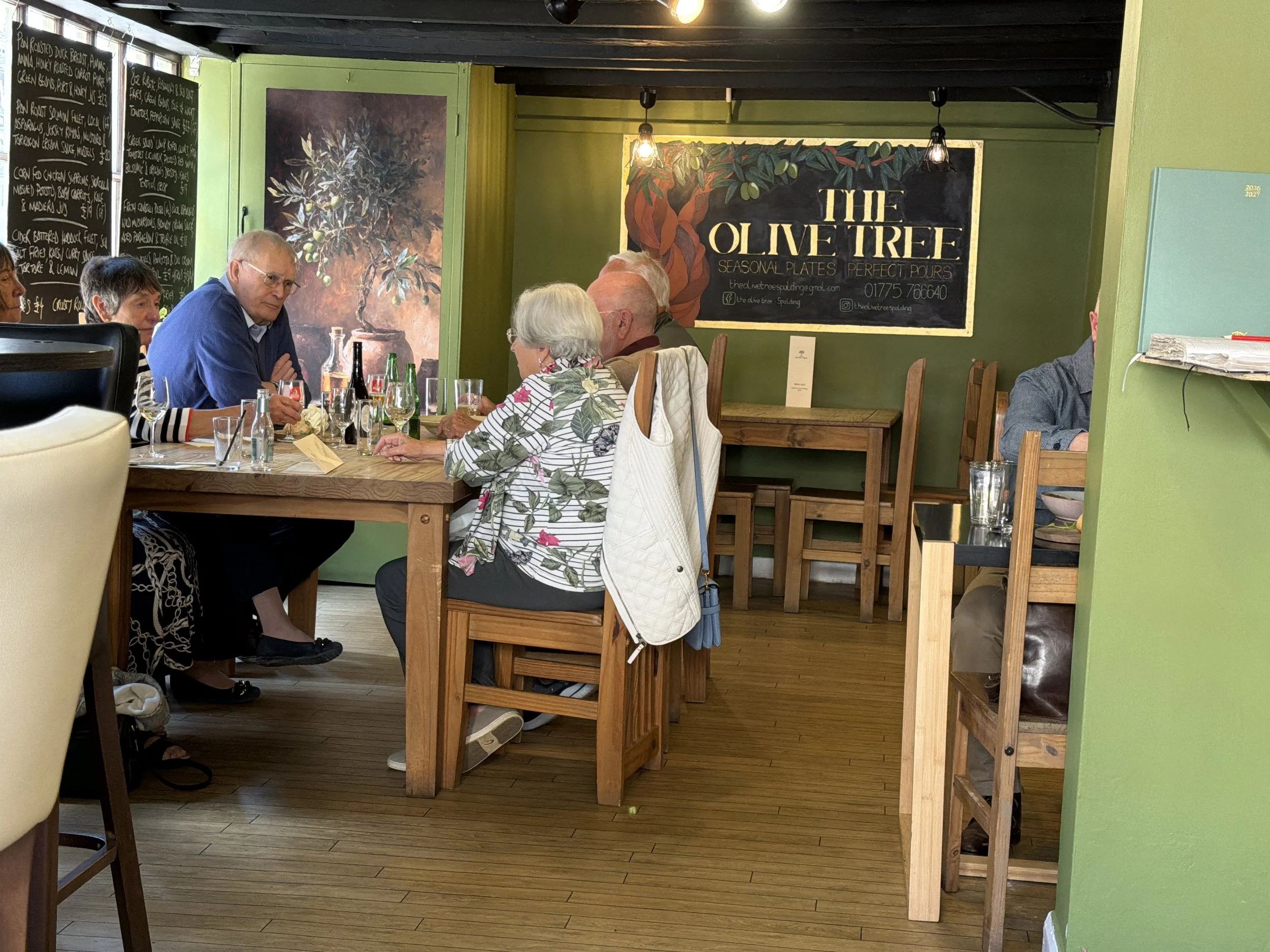 A group of older adults sitting at a long wooden table inside a cozy restaurant named 'The Olive Tree'. The restaurant has green walls, decorative artwork of trees, and chalkboard menus on the walls. The patrons are engaged in conversation, and the t