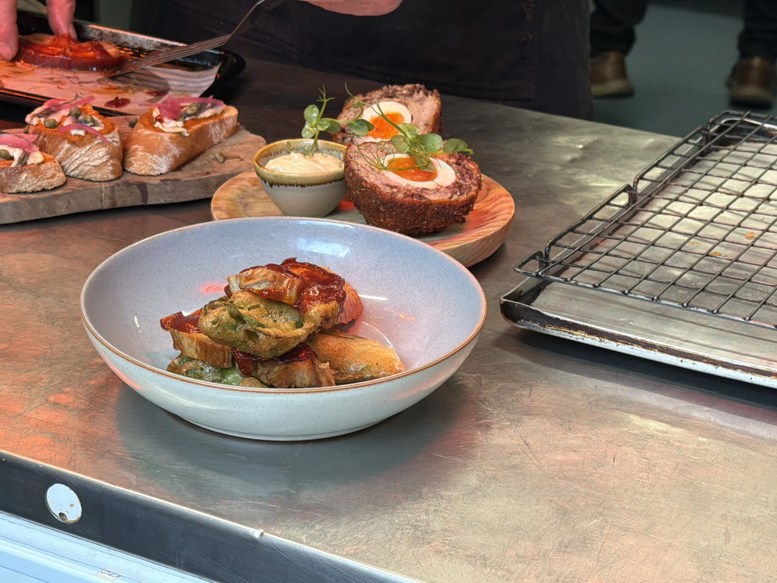 Close-up of a kitchen counter with prepared food including a sliced meatloaf with a soft boiled egg inside, garnished with herbs, served on a wooden platter. There is also a small bowl of sauce and a ceramic plate with layered vegetable and meat dish