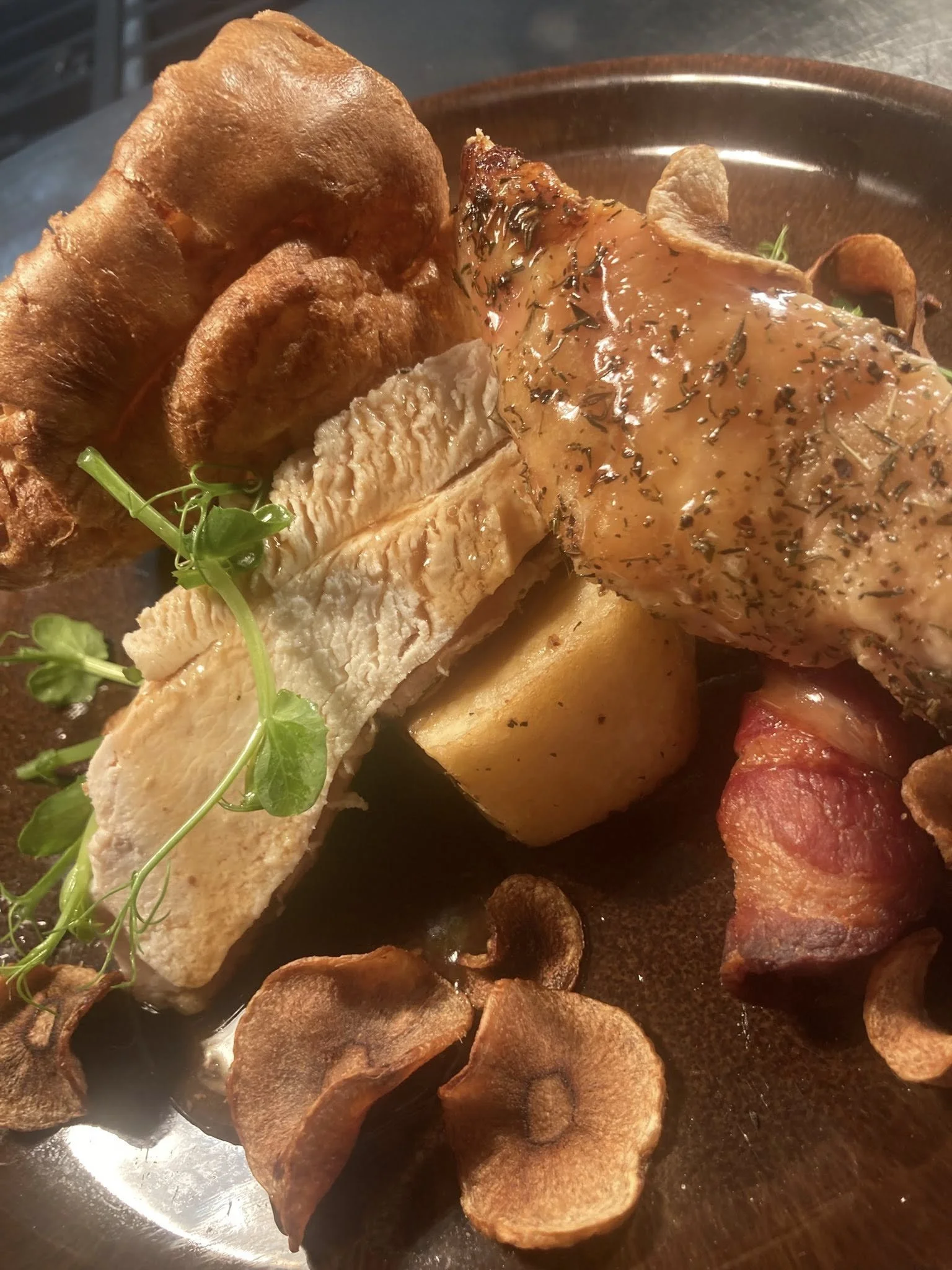 Roasted chicken breast with herbs, served with crispy potato slices and garnished with fresh greens, all on a dark plate.