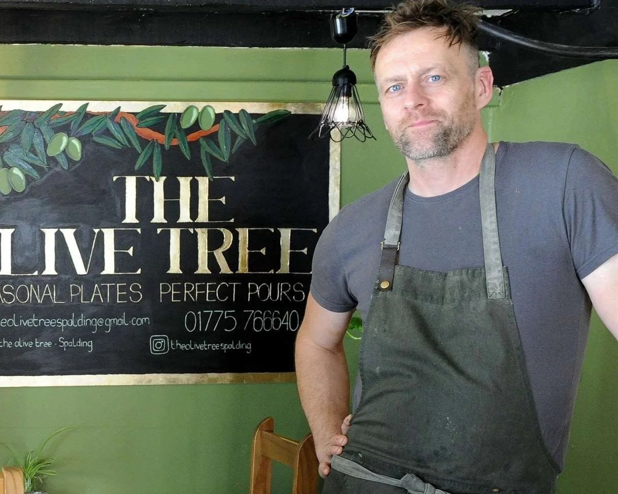 A man with a beard and short hair wearing a dark gray t-shirt and black apron standing in front of a blackboard sign that reads "The Olive Tree" and offers personal plates. The sign has a painted olive branch and contact information. The background features green walls and a light fixture hanging from the ceiling.