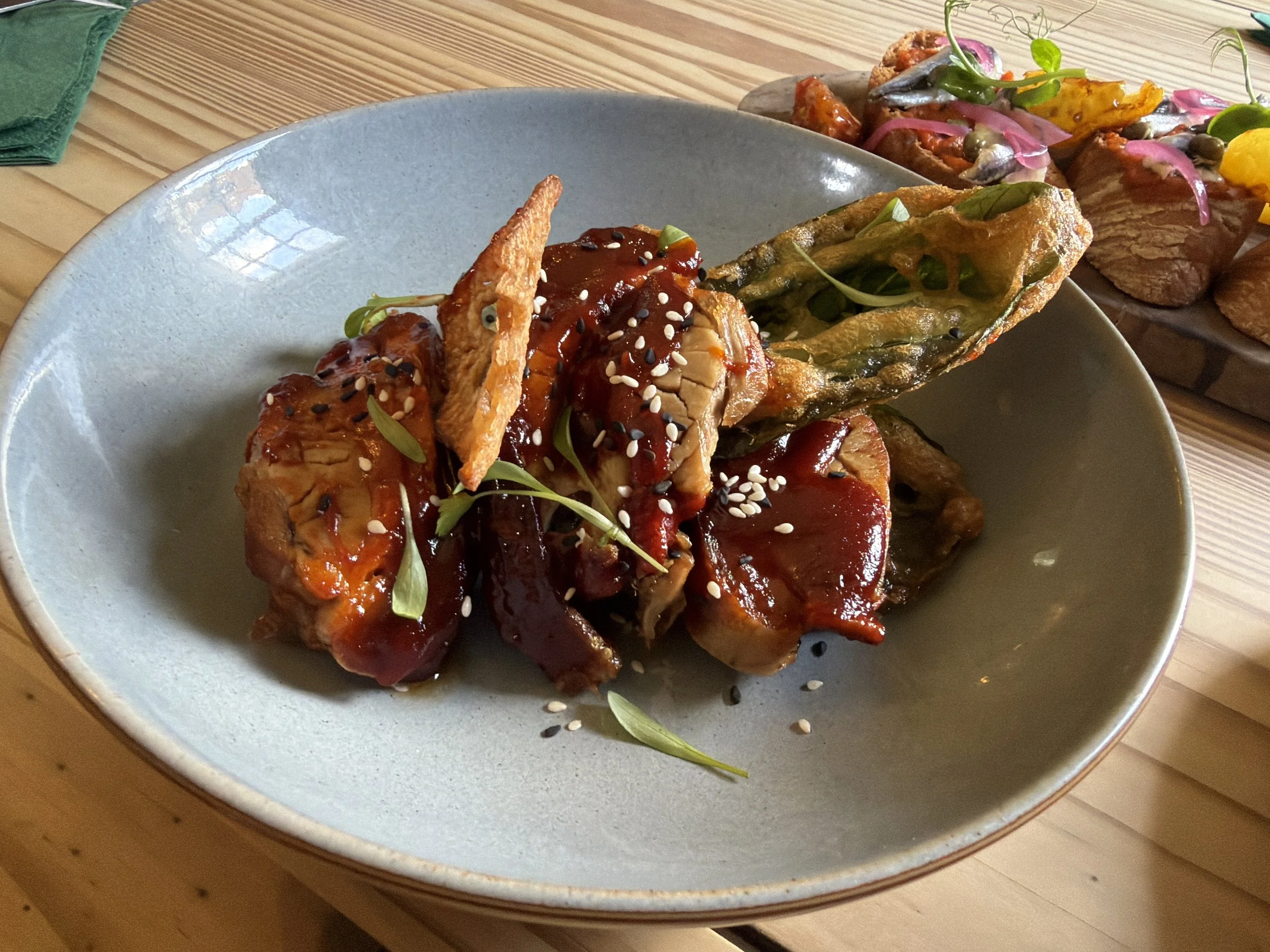 A ceramic bowl with glazed interior containing Asian-style grilled chicken wings topped with sauce and garnished with sesame seeds and microgreens. In the background, there is a plate of stuffed peppers with sauce and garnishes on a wooden table.