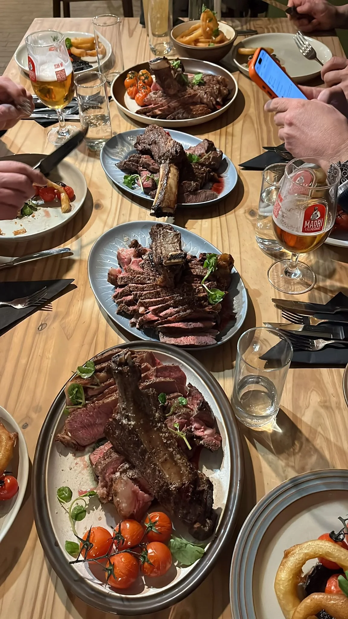 A table filled with various plates of cooked meats, including steak and ribs, surrounded by glasses of beer and water. There are also bowls of side dishes such as fries and salads, and people are seen taking photos and eating.