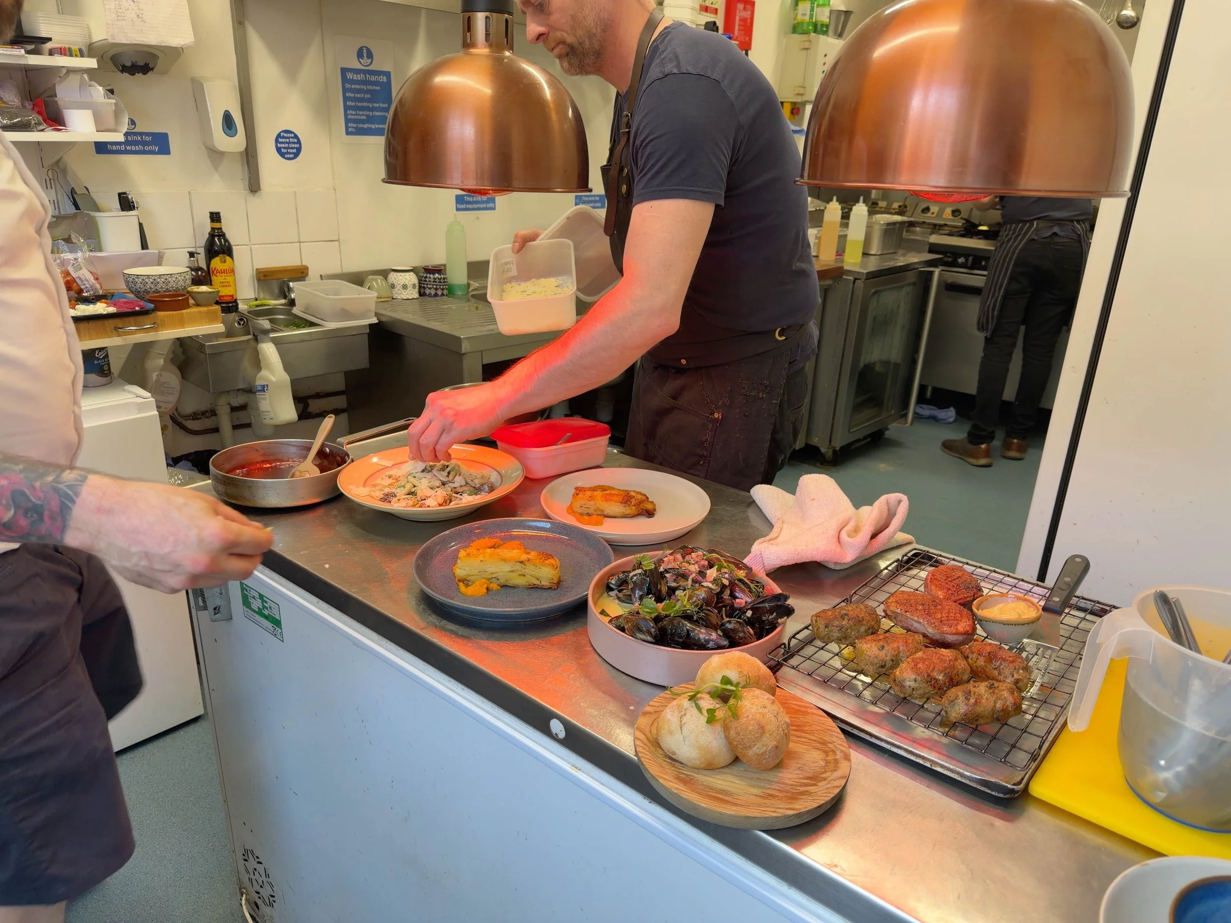 A person in a kitchen preparing food on a metal countertop with various dishes, including bread rolls, baked goods, salads, and cooked items. There are copper heat lamps hanging above, and the background has kitchen supplies and cooking utensils.