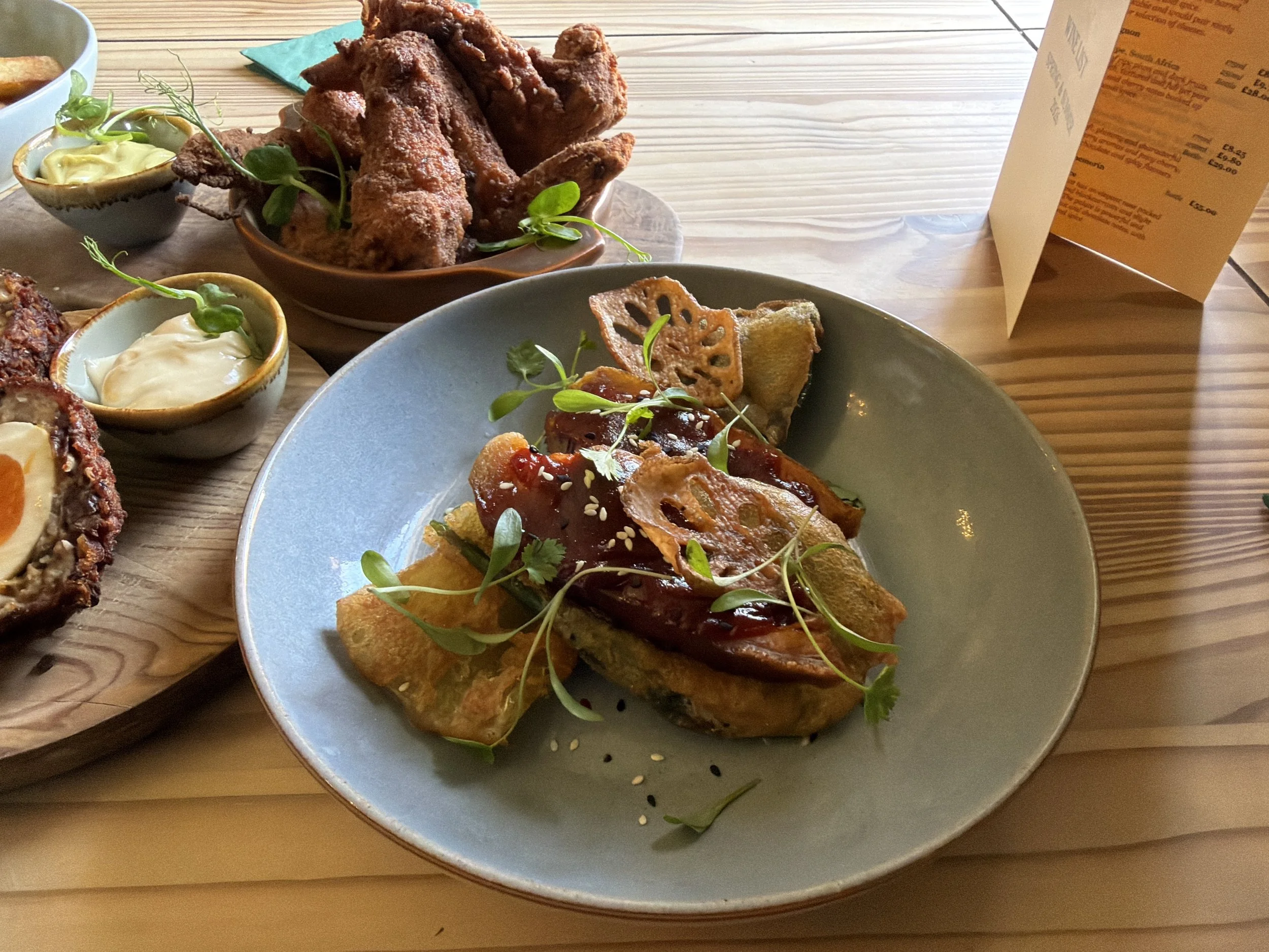 A plate of Asian-inspired appetizer with grilled vegetables and microgreens, featuring a lotus root chip, topped with a glaze and sesame seeds.