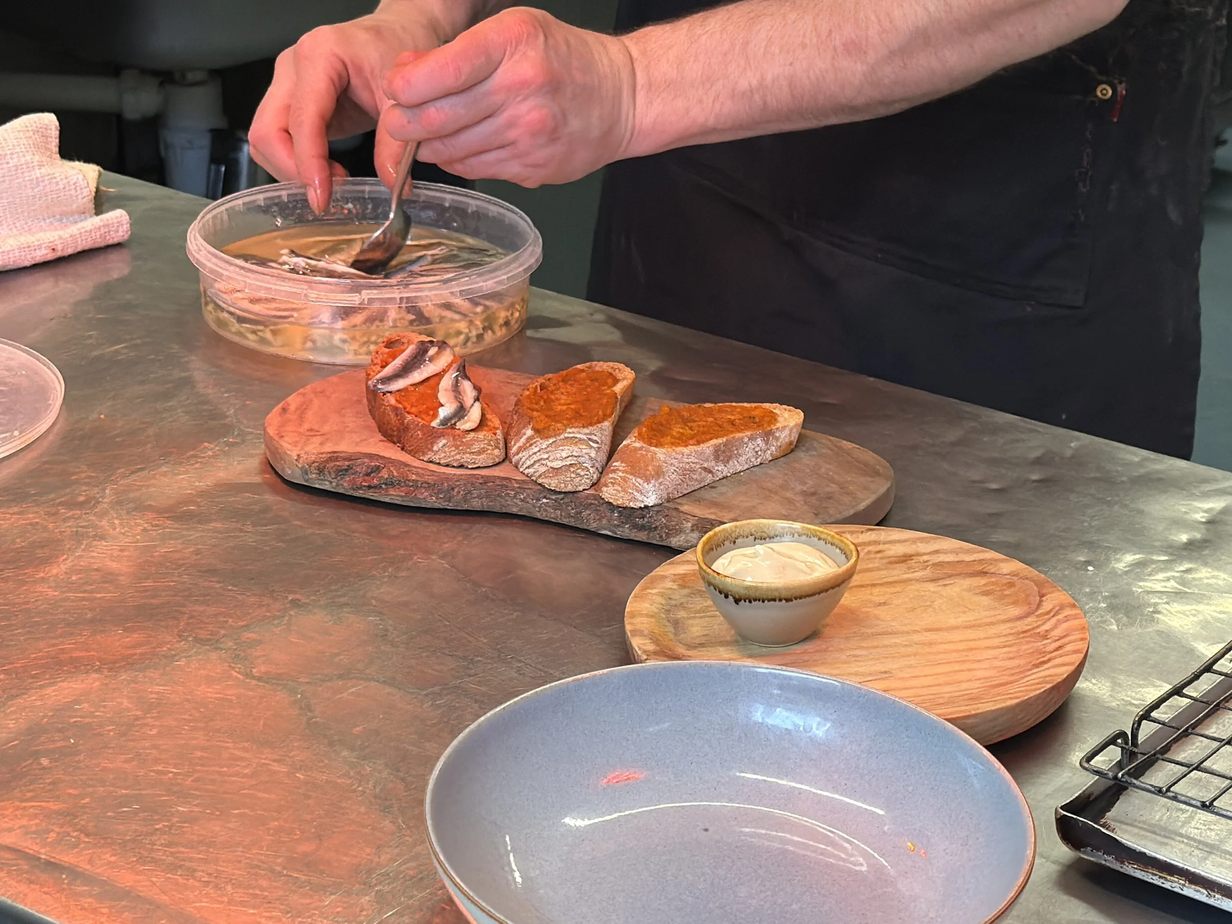 Person assembling hot dog toppings on a wooden board with bread, fish fillets, and condiments nearby.