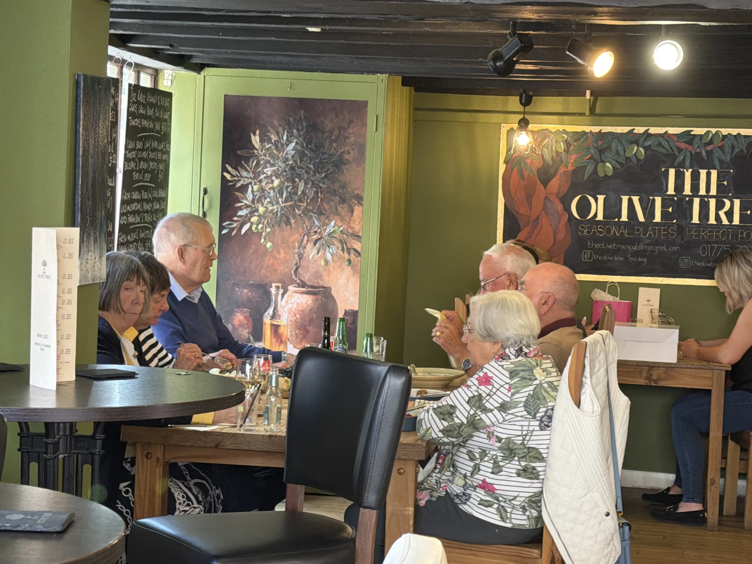 People dining at a restaurant, sitting at wooden tables with glasses and bottles, decorated with paintings and green walls.