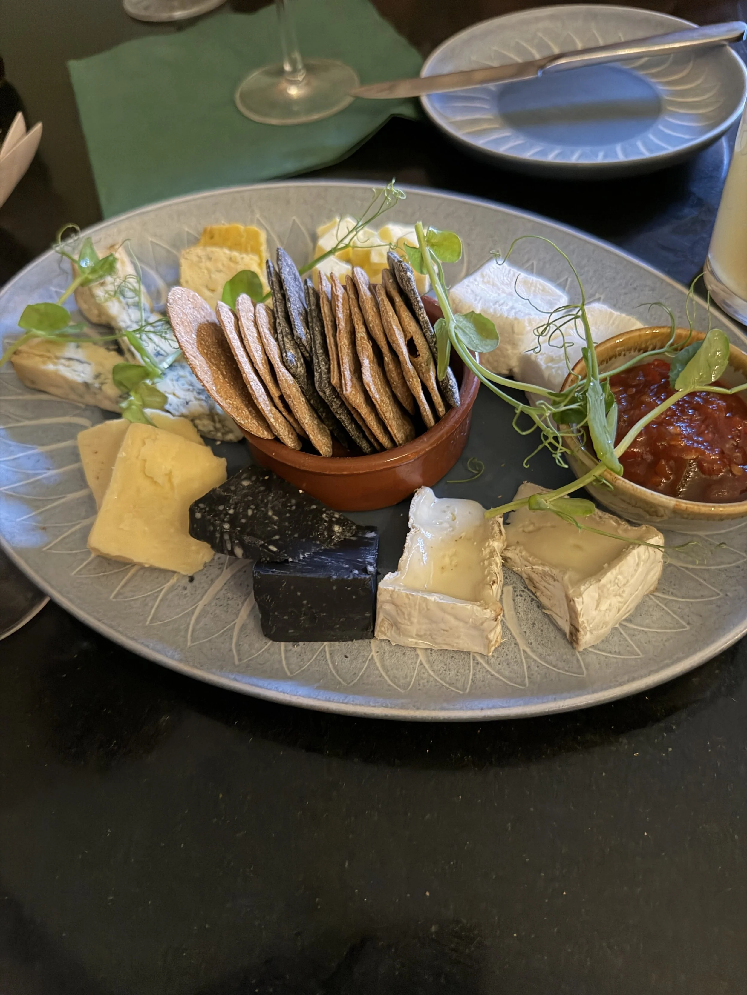 A charcuterie platter with various cheeses, crackers, and a small bowl of jam on a large gray plate, garnished with fresh green herbs. There is a glass of wine, a small empty plate with a knife, and a green napkin in the background.