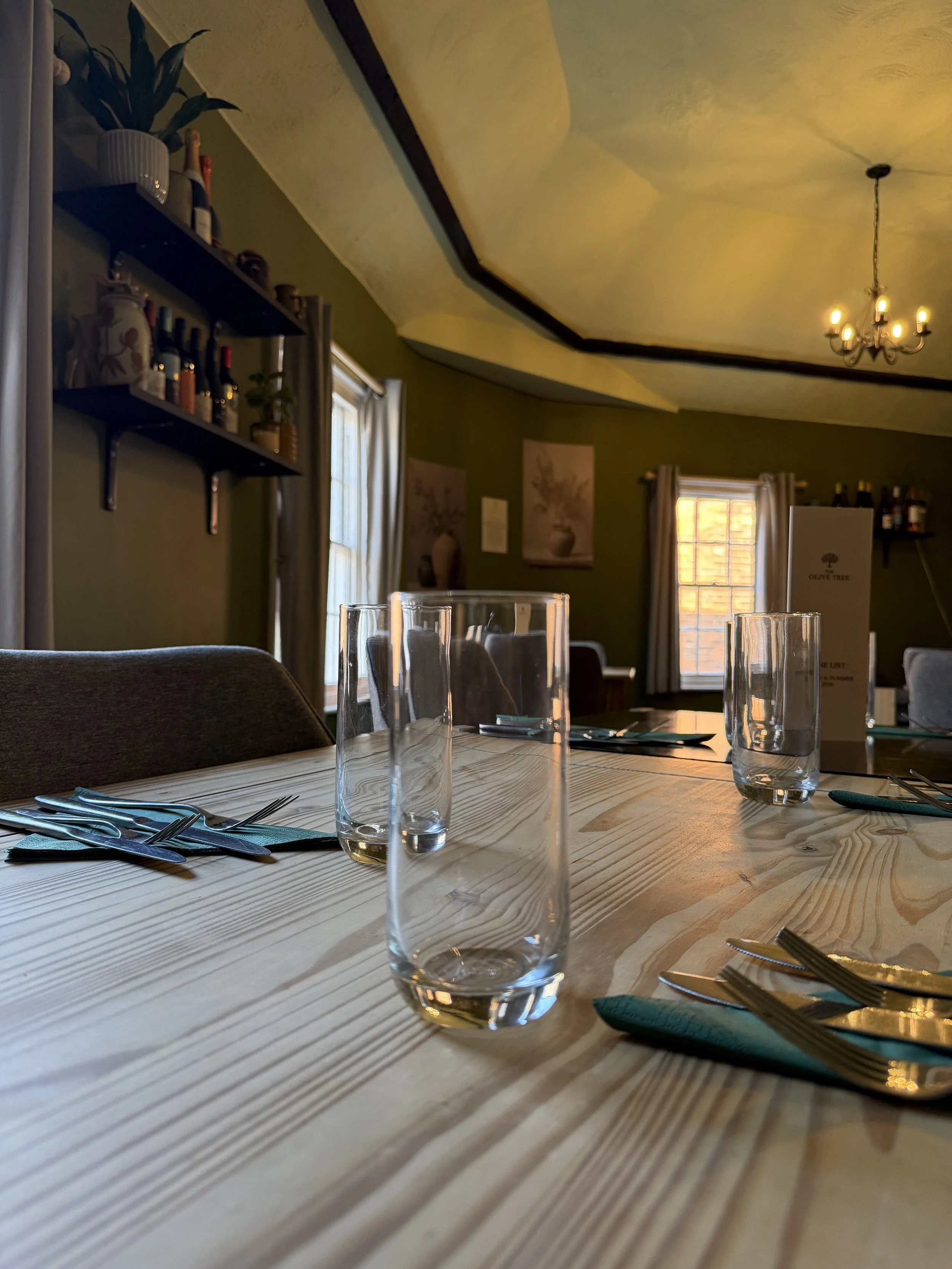 A dining table set with drinking glasses, forks, and teal cloth napkins, in a warmly lit room with sunlight coming through windows and a chandelier hanging from the ceiling.