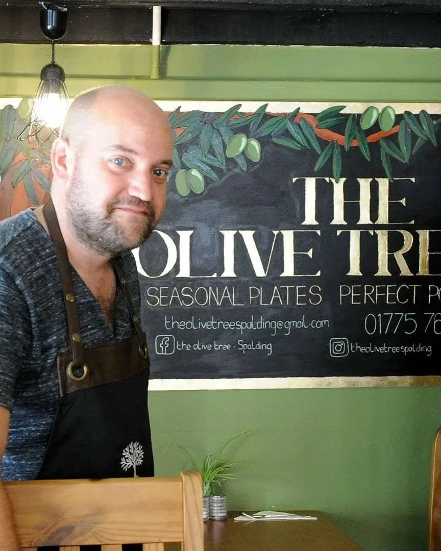 A man with a beard and a shaved head standing in front of a chalkboard menu inside a restaurant named The Olive Tree. The menu mentions seasonal plates and that they offer perfect P. The background features green walls and hanging light fixtures.