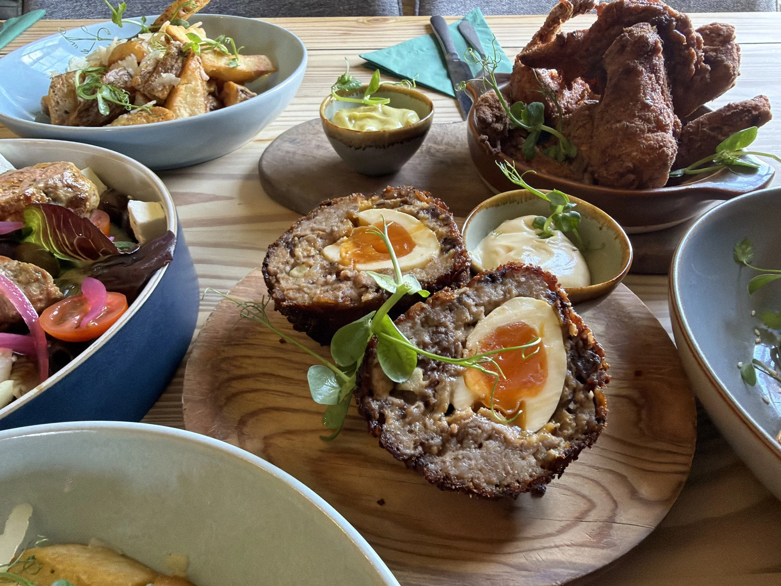 A variety of food dishes on a wooden table, including meatloaf with a baked egg on top, a bowl of fried chicken drumsticks, a bowl of roasted sweet potato wedges, small bowls of dipping sauces, and side salads with vegetables and sprouts.