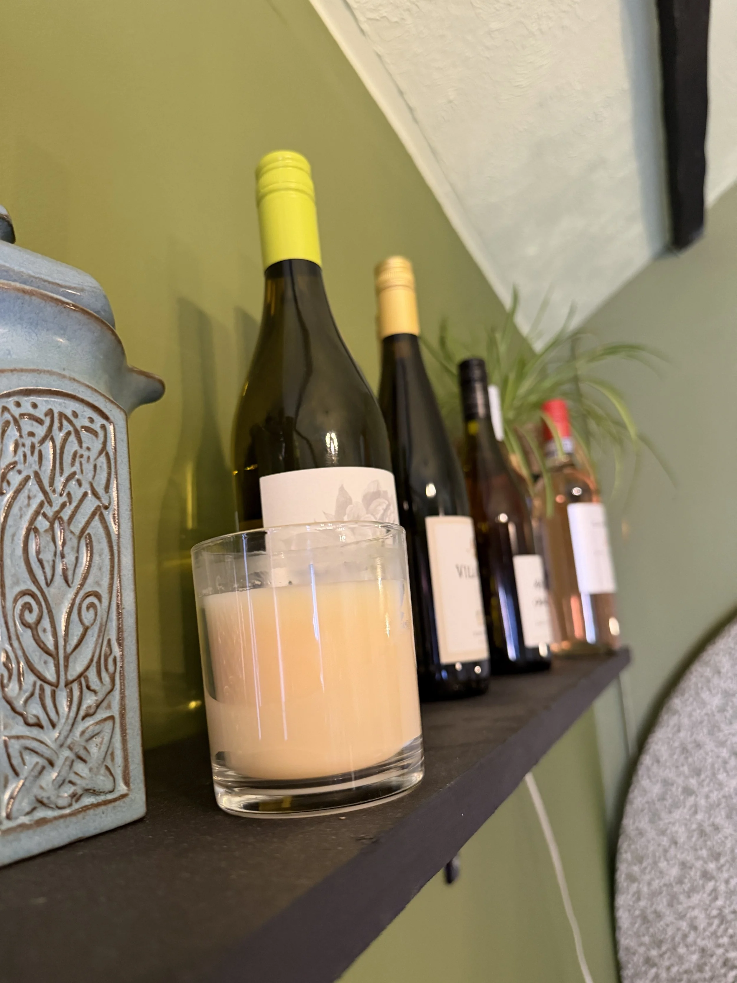 Close-up of a black shelf with several bottles of wine, a lit candle, a decorative metal container, and a small plant against a green wall.