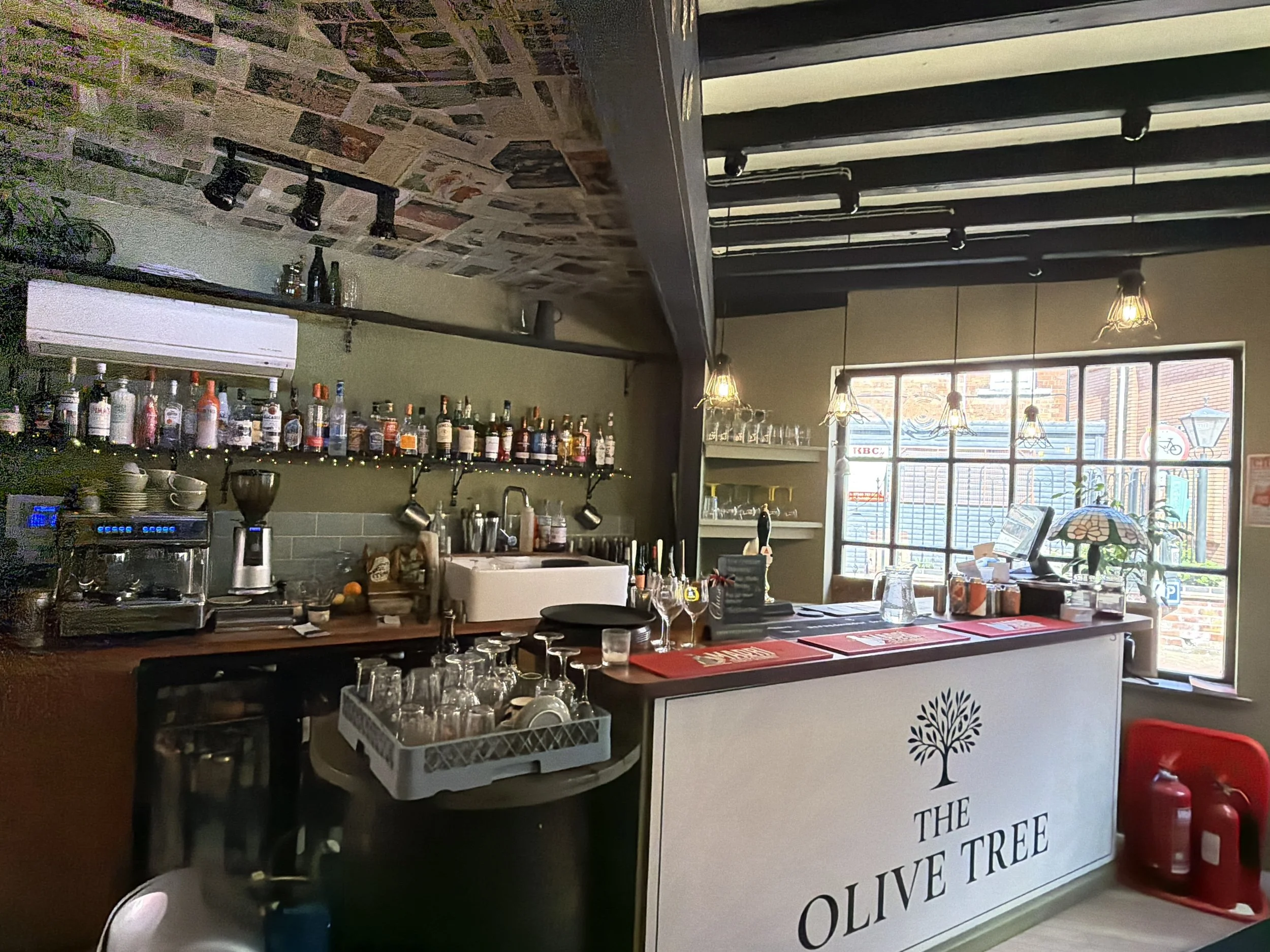 Interior of a cafe or bar with shelves of bottles, hanging lights, and a window showing the outside street. A counter with glasses and a sign that reads 'The Olive Tree' and a fire extinguisher nearby.
