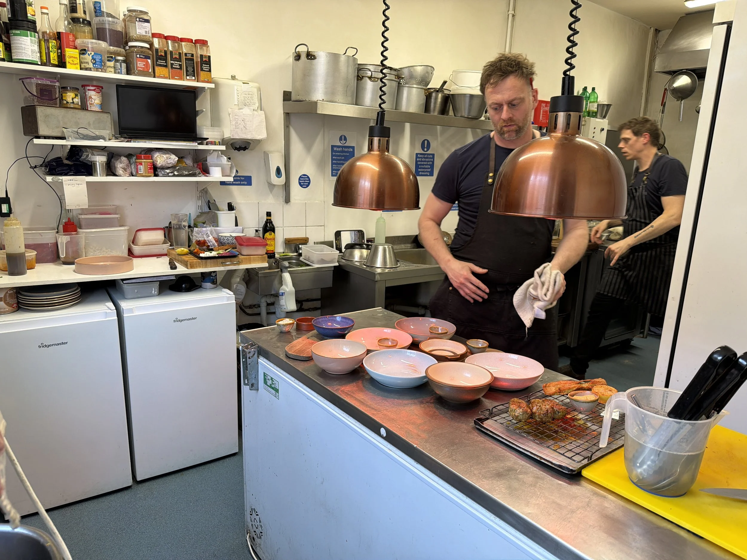 A busy commercial kitchen with two male staff members preparing plated dishes. The kitchen is equipped with shelves holding spices, containers, and kitchen supplies. There are copper pendant lights hanging over the workspace, with various bowls and f