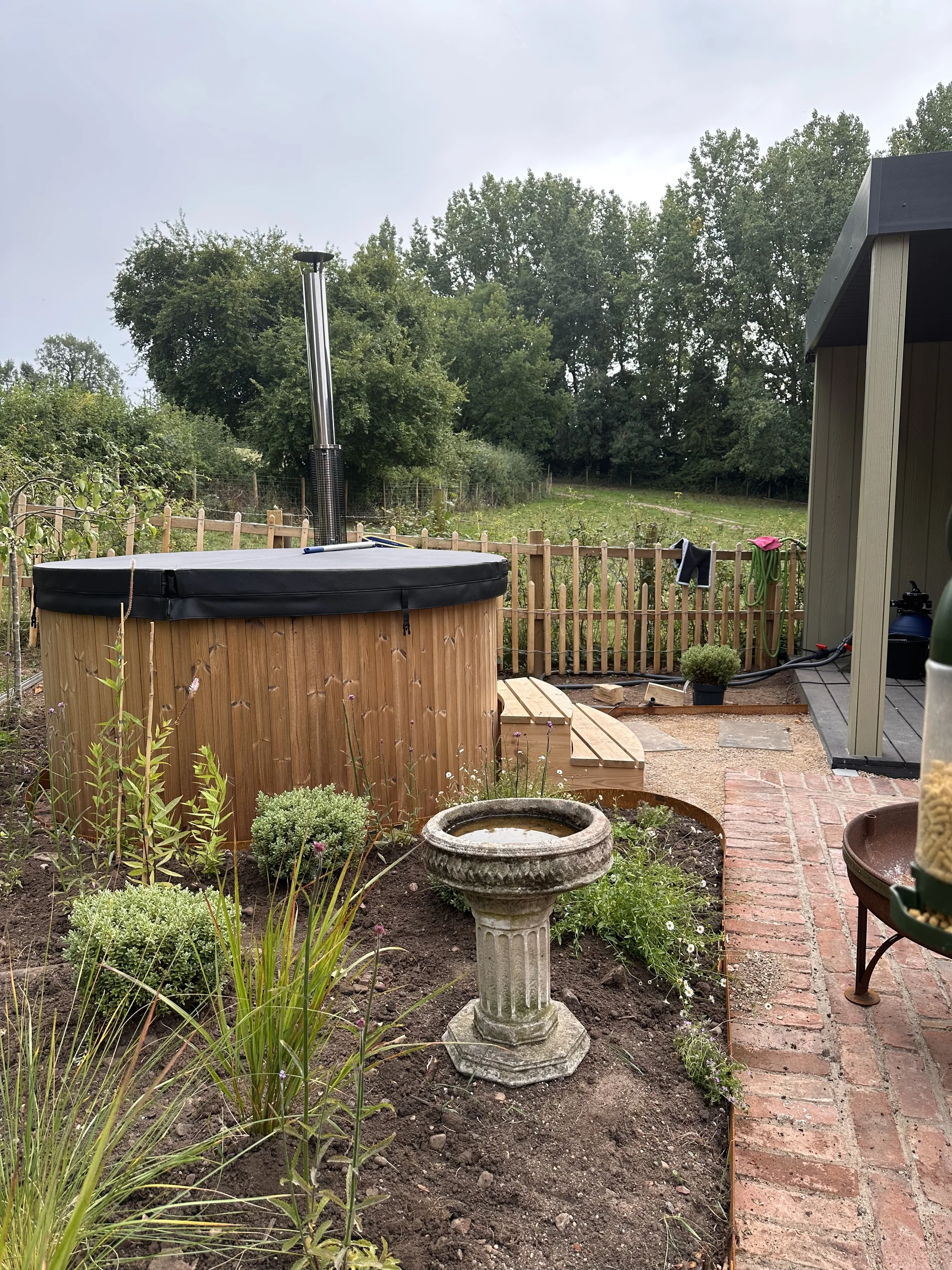 Backyard with hot tub, bird bath, garden, potted plant, brick and wood patio, fence, and trees.