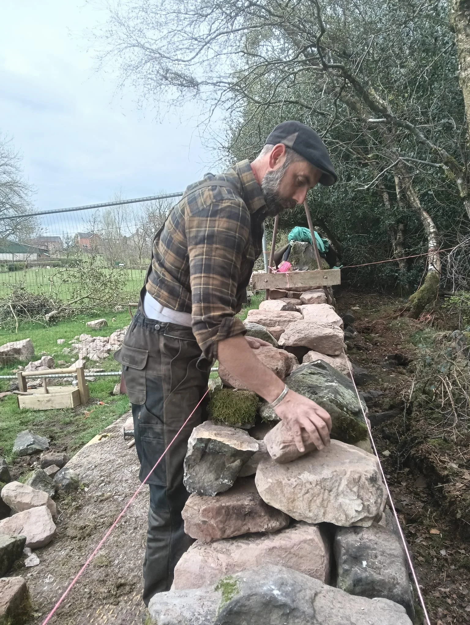 A man stacking large rocks to build a dry stone wall outdoors, with trees and a field in the background.
