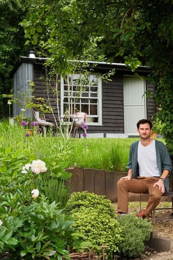 A man sitting on a chair in a lush garden with green plants, flowers, and a small dark-colored shed in the background.