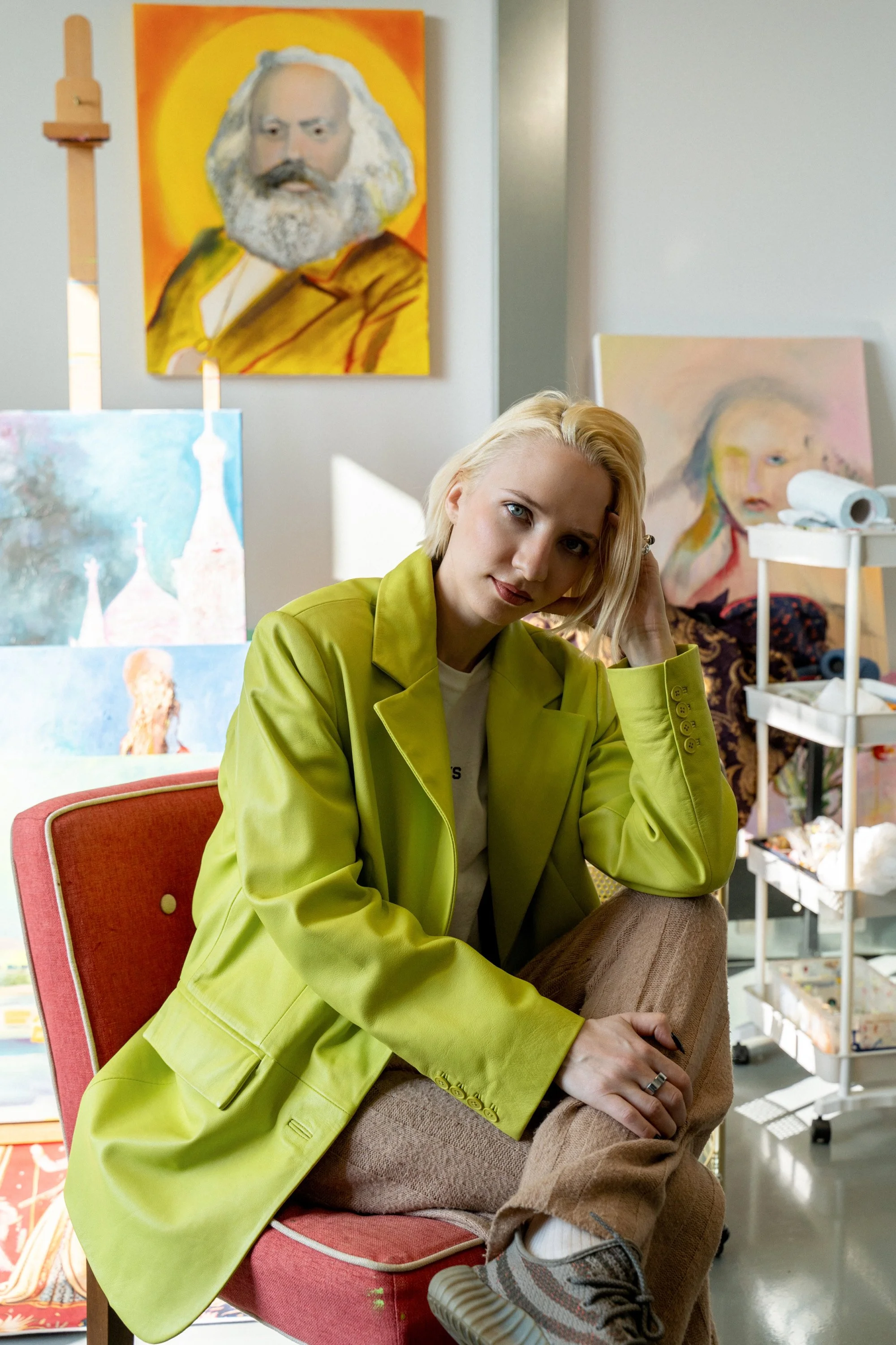 A young woman with blonde hair and blue eyes sitting on a pink chair, wearing a light green blazer and beige pants, with her hand on her head, in an art studio with paintings and art supplies in the background.