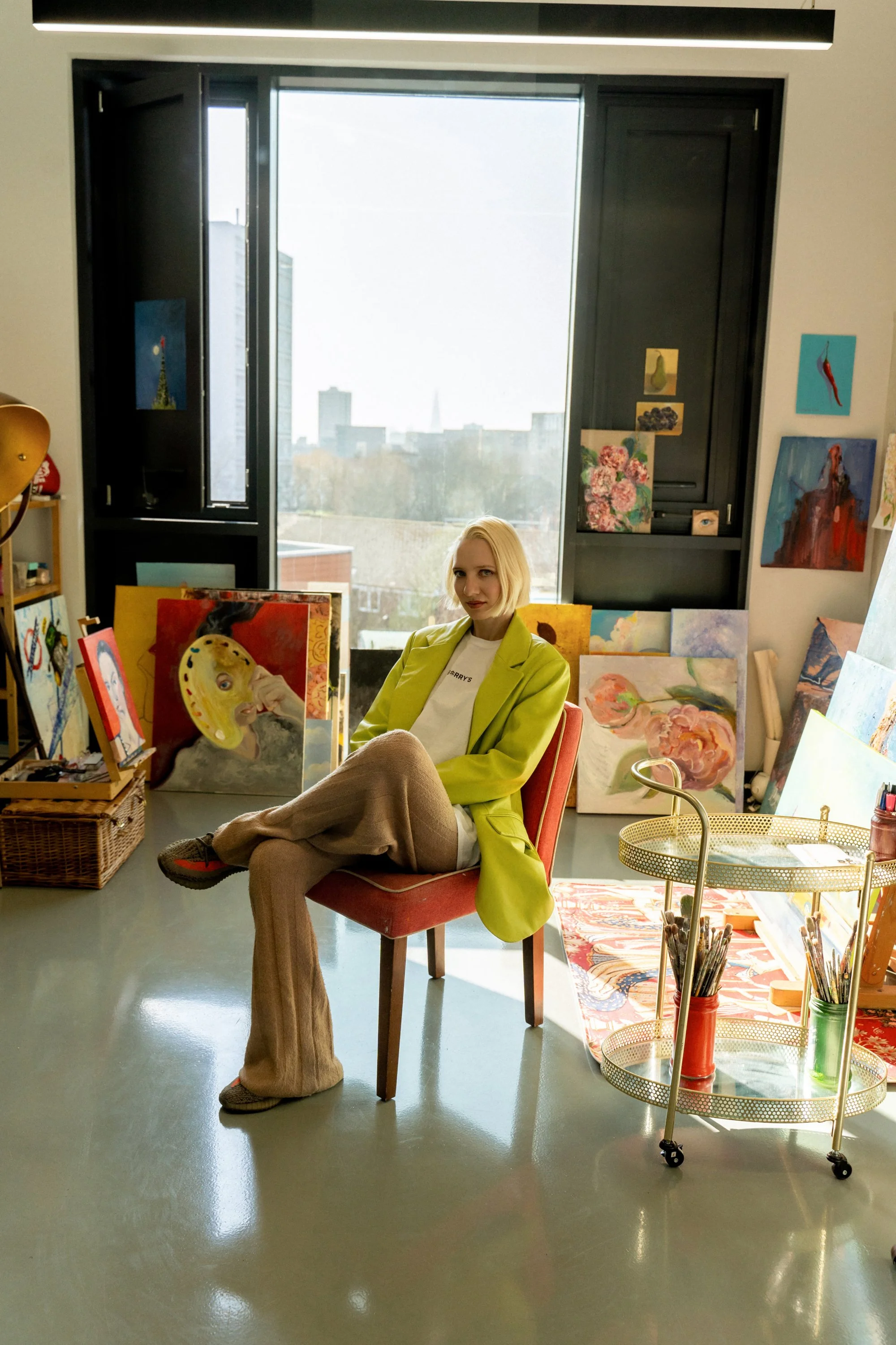 A woman with blonde hair wearing a lime green blazer and beige wide-leg pants sitting on a red chair in an art studio.